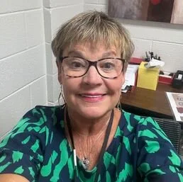 A smiling woman with short blonde hair and glasses, wearing a green patterned top, in an indoor setting with a desk and office supplies in the background.