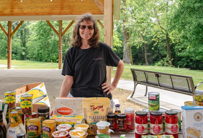 Woman smiling at a vendor table with canned and jarred food products outdoors under a pavilion, with trees and a park bench in the background.