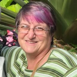 Smiling woman with short hair dyed purple and pink, wearing glasses and a green striped shirt, in a garden with green foliage and pink flowers.