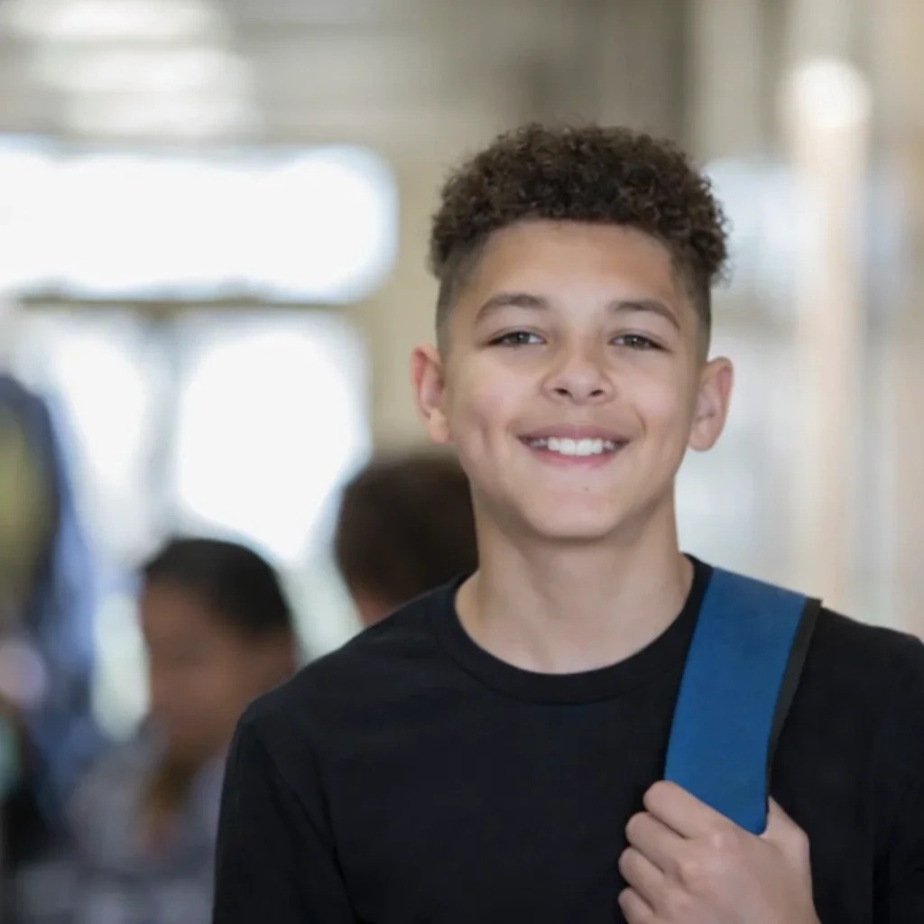 A smiling young boy with curly hair wearing a gray shirt and a blue backpack in a school hallway.