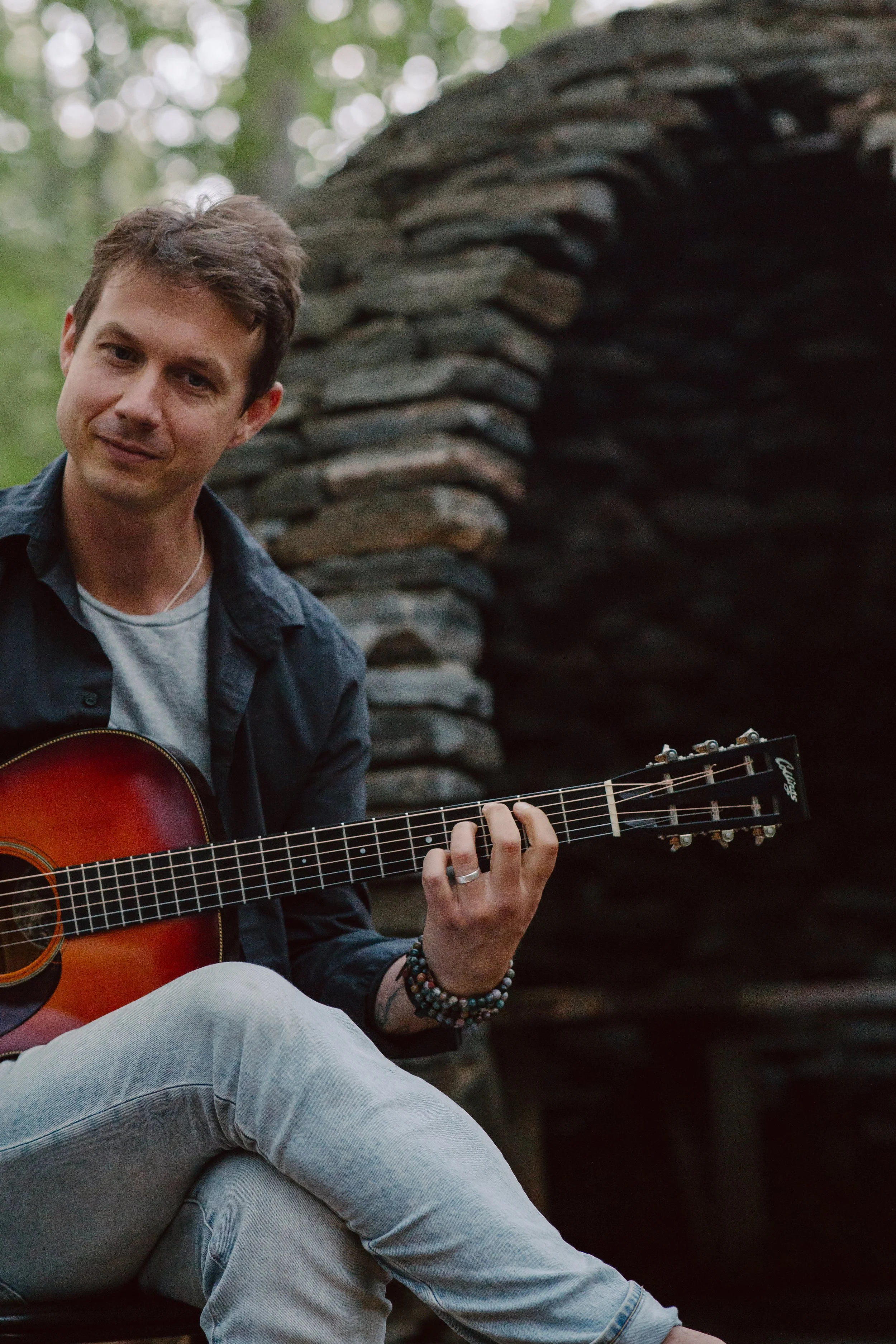A young man sitting outdoors playing an acoustic guitar, with a blurred stone structure behind him and green trees overhead.