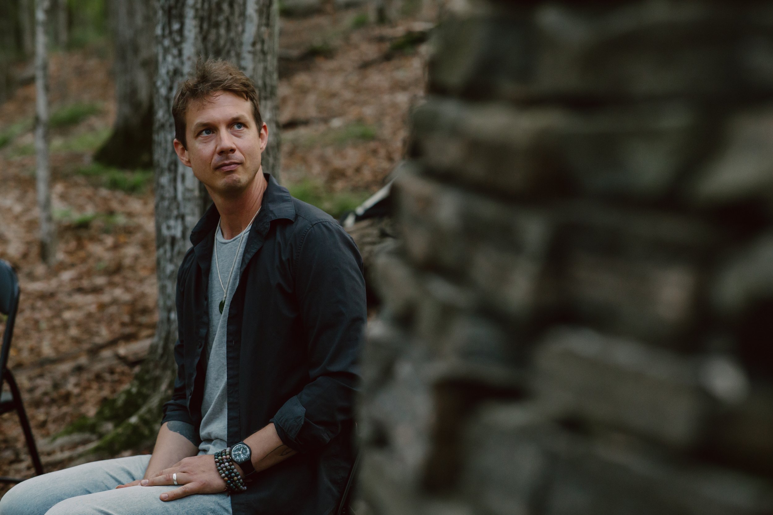 A young man with light brown hair, blue eyes, wearing a dark jacket, a gray t-shirt, and multiple beaded bracelets sits on a log in a forested area, looking to the side.