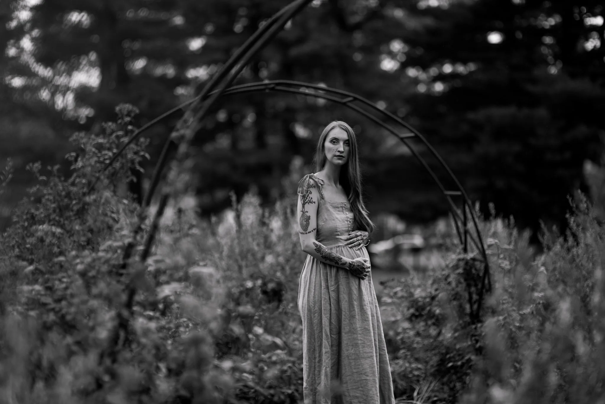 A young woman with long hair and tattoos standing in a natural outdoor setting, framed by an arched metal structure, in black and white photography.