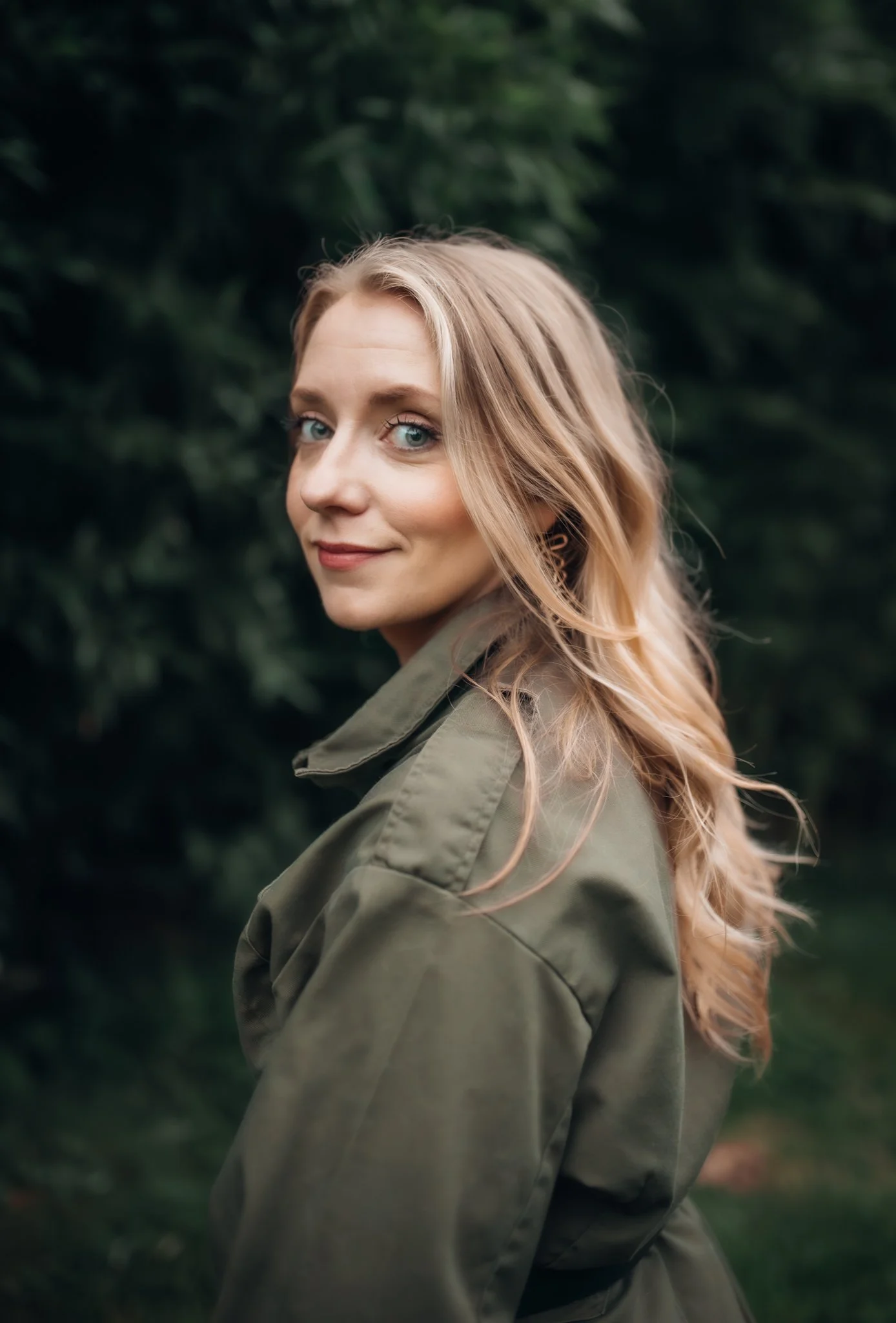 A woman with blonde wavy hair and blue eyes wearing an olive green jacket outdoors with dark green leafy background.