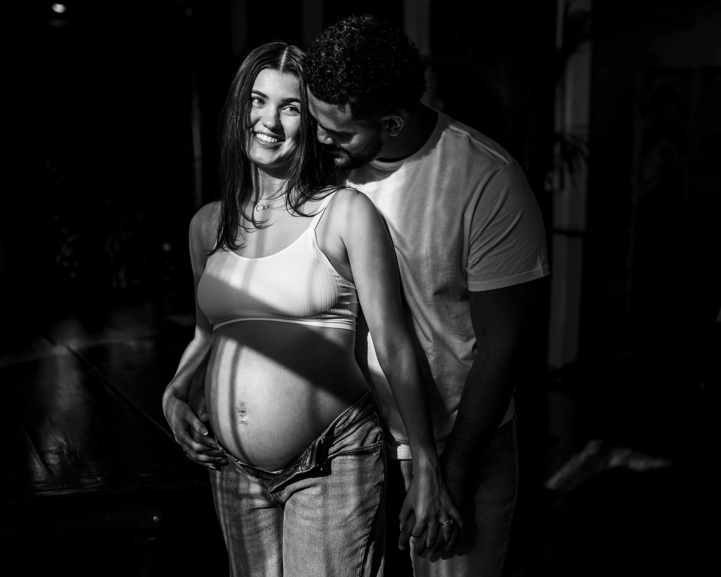 A black and white photo of a pregnant woman smiling while her partner stands close to her, touching her belly and holding her hand in a dimly lit room.