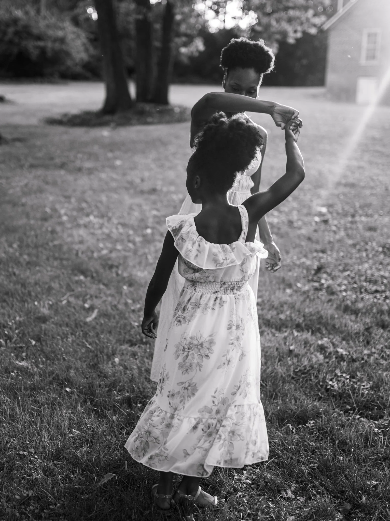 A woman and a young girl dancing outdoors on a grassy area, with sun rays and trees in the background, captured in black and white.
