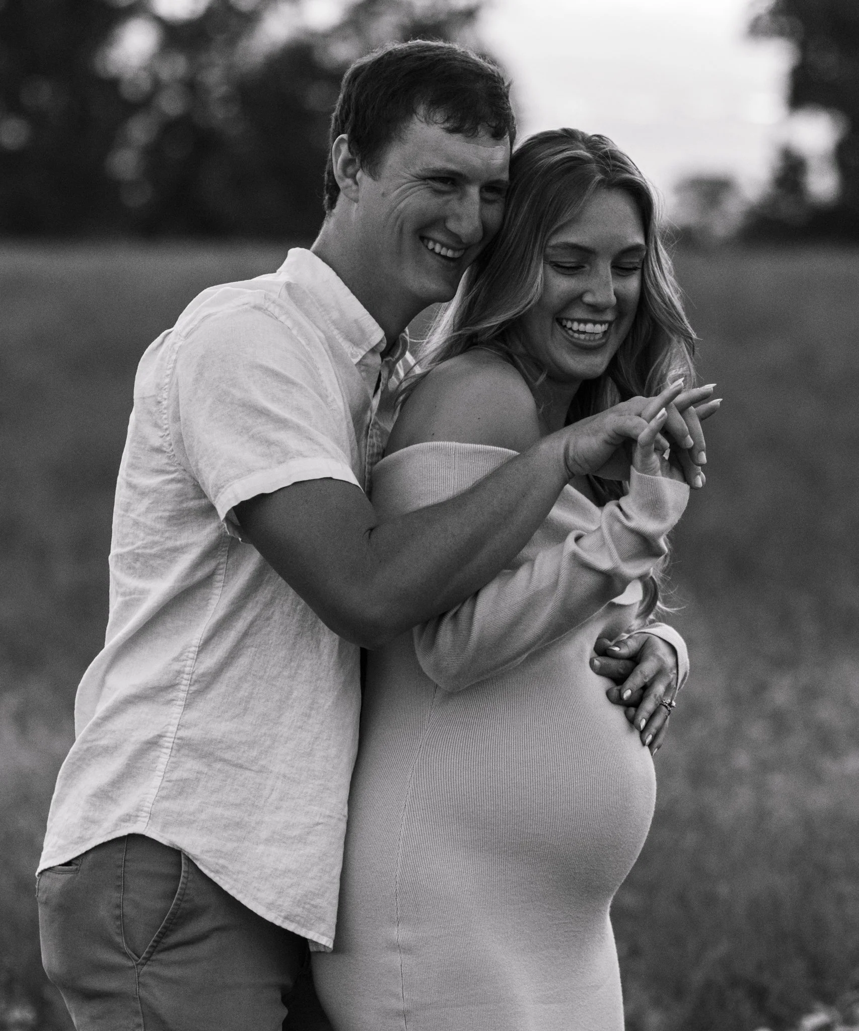 A smiling pregnant woman and a man are outdoors. The man embraces the woman from behind, both are looking down and touching her baby bump with their hands.