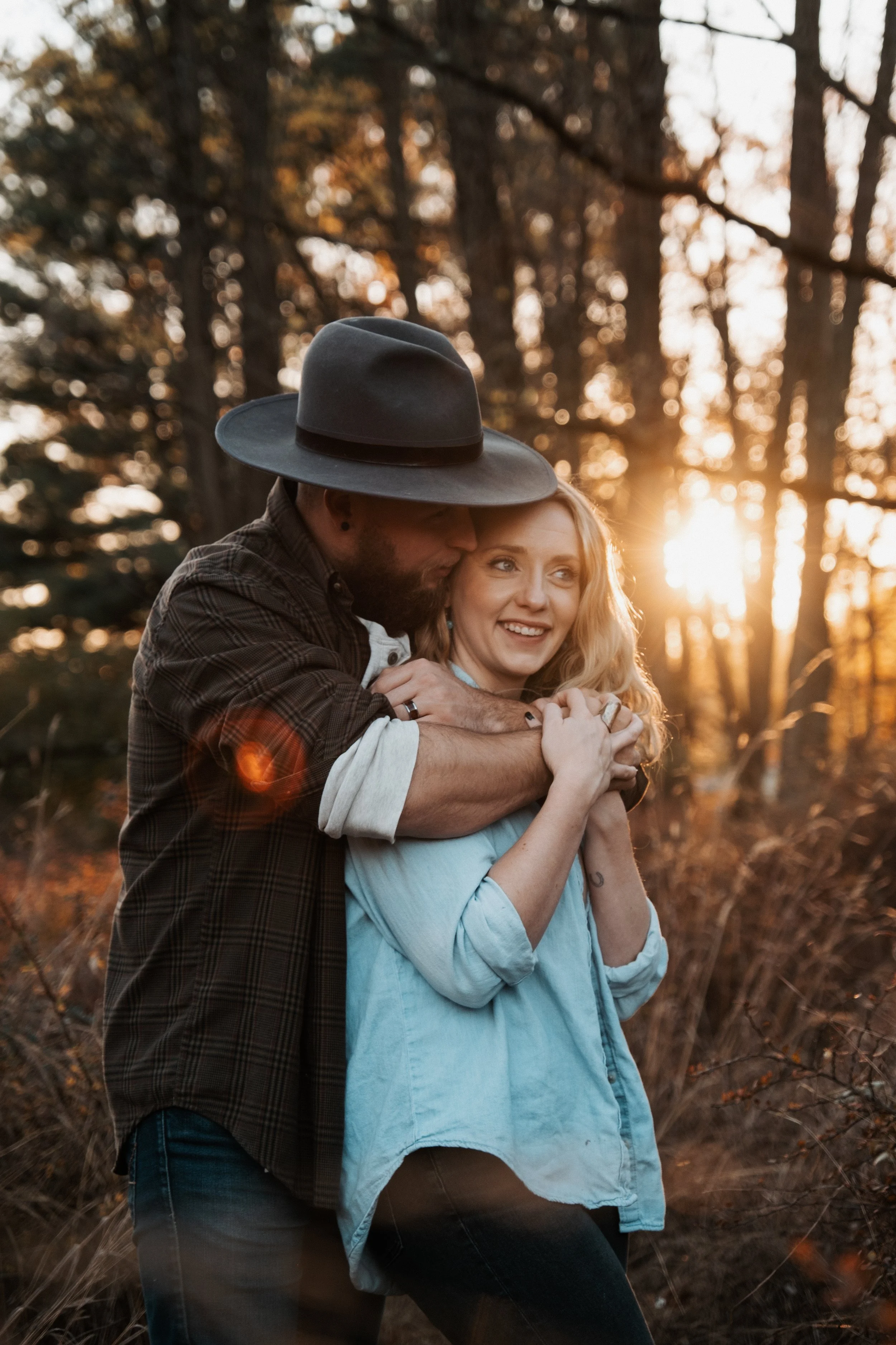 A man with a beard wearing a wide-brimmed hat and casual clothing is hugging a woman with wavy, blonde hair dressed in a light blue shirt. They are outdoors with trees and the setting sun in the background.