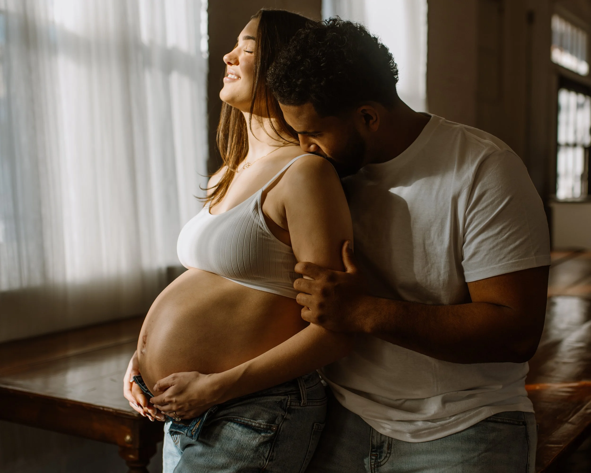 a pregnant woman rests against her husband's chest as he kisses her shoulderin their cozy studio maternity session in Bethlehem PA