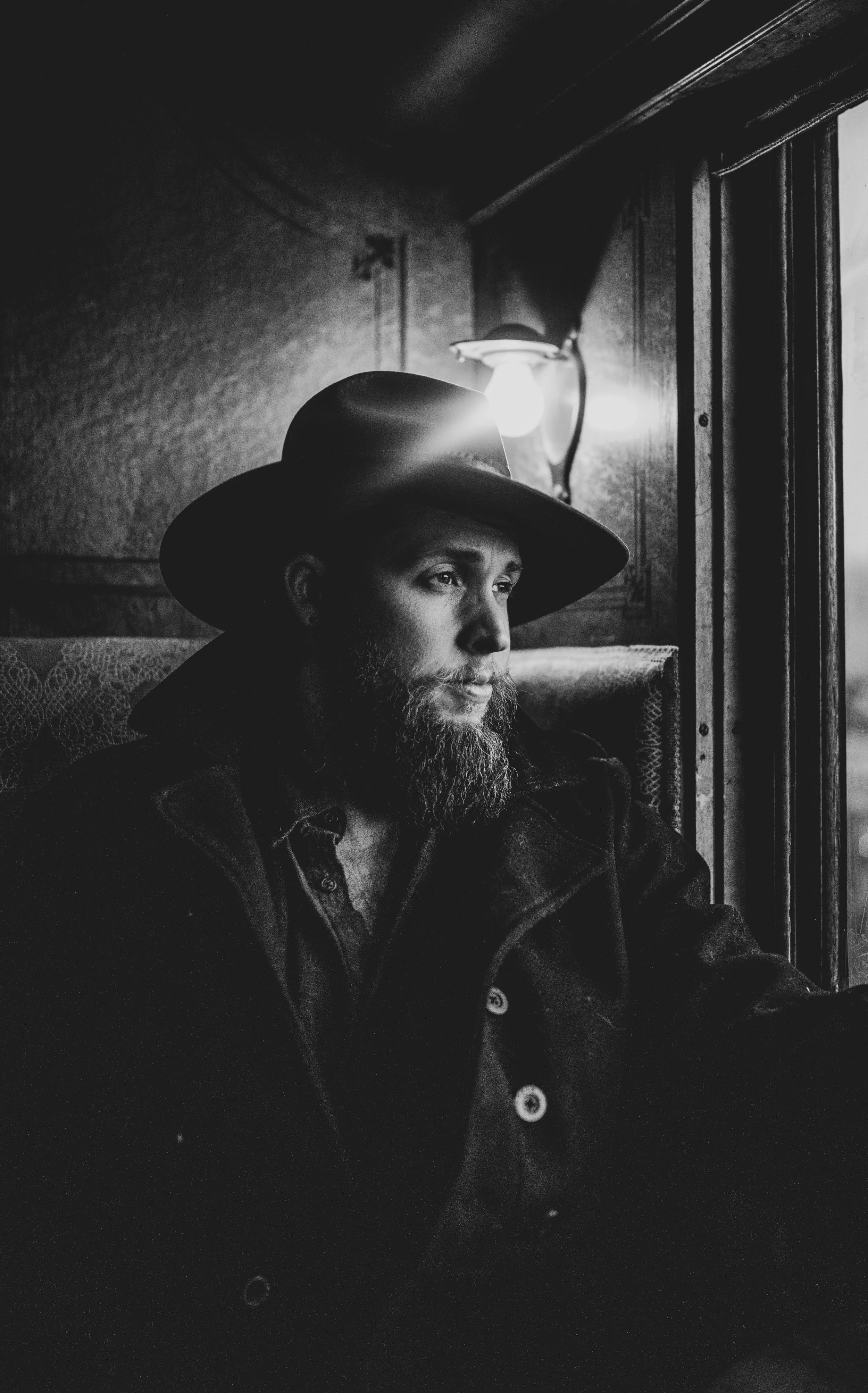 Black and white photo of a man with a beard wearing a wide-brimmed hat, sitting and looking out of a window inside a train.