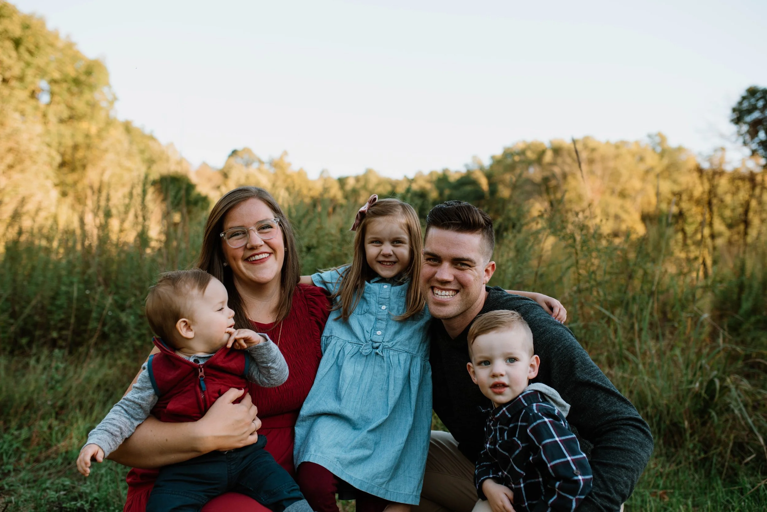 A smiling family of five outdoors in a grassy field with trees in the background.