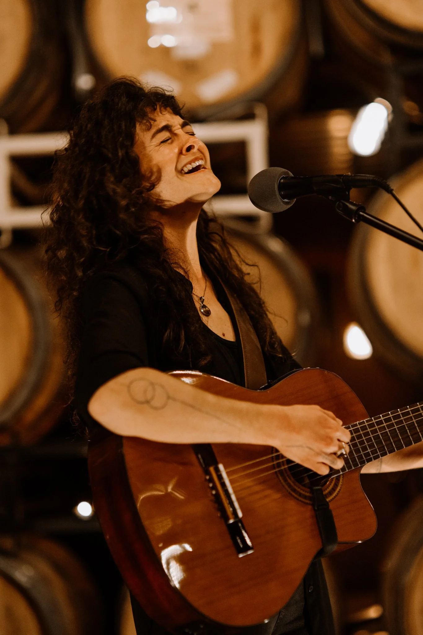 A woman with curly hair singing passionately while playing an acoustic guitar in front of a microphone, with wooden barrels in the background.
