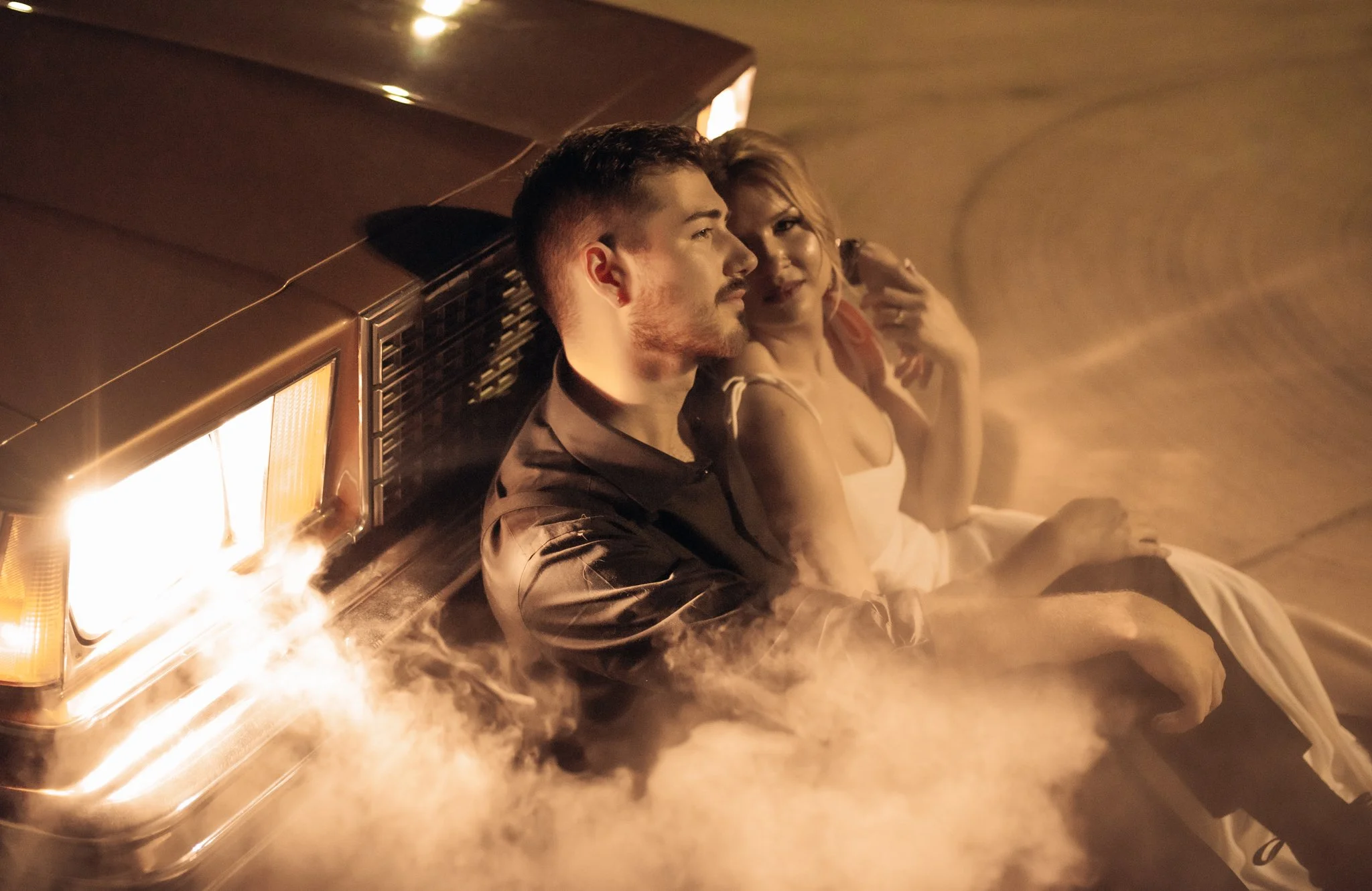 A man and woman sit on the ground next to a vintage car with smoke surrounding them in a moody, dimly lit scene.