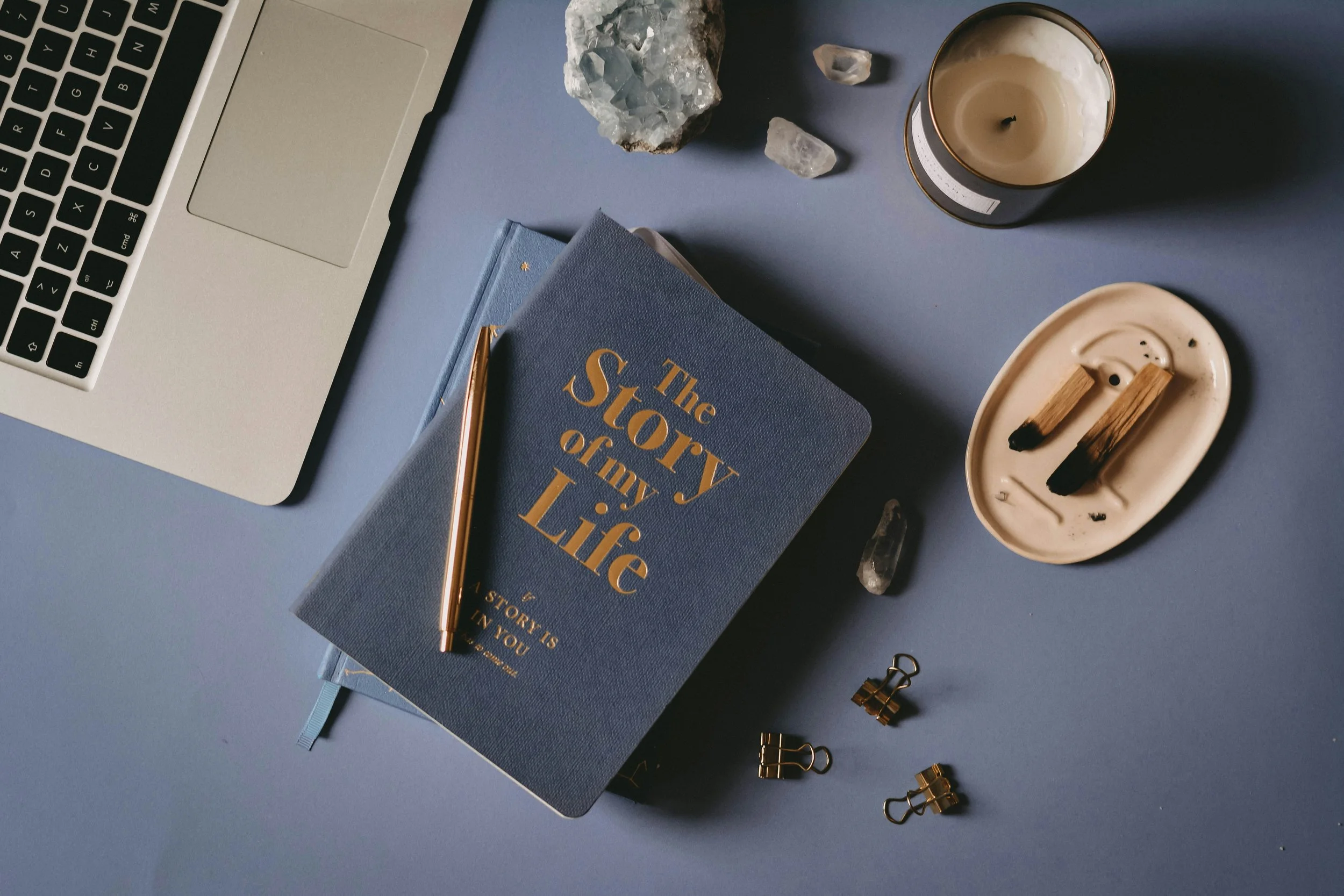 Journal and book on top of a table with a golden pen, open laptop, candle, and other desktop items.