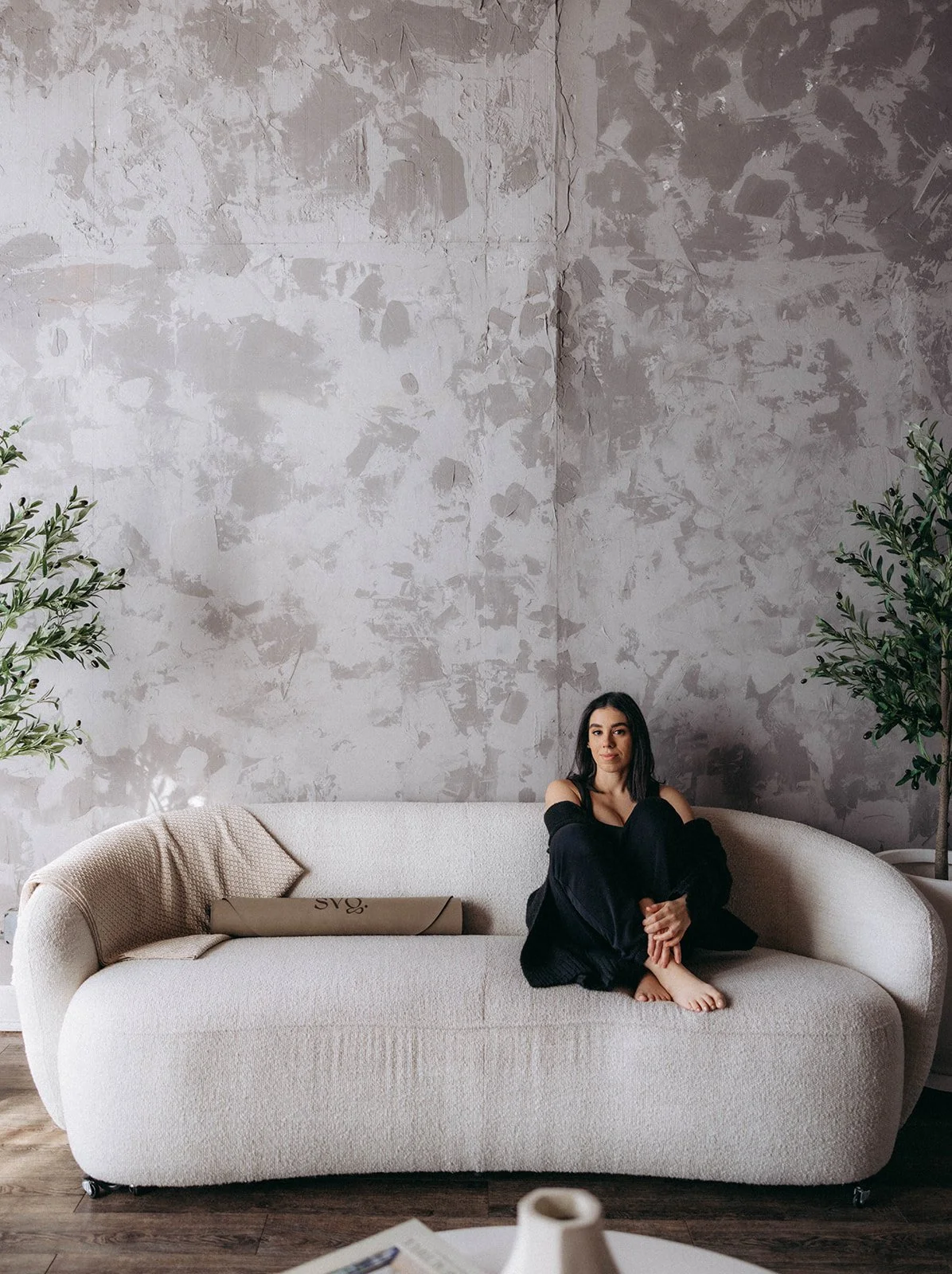 A woman dressed in black is seated on a white luxe couch with the Leche de Perla Capsula Mat rolled up and placed on the other side of her with an aesthetic backdrop.