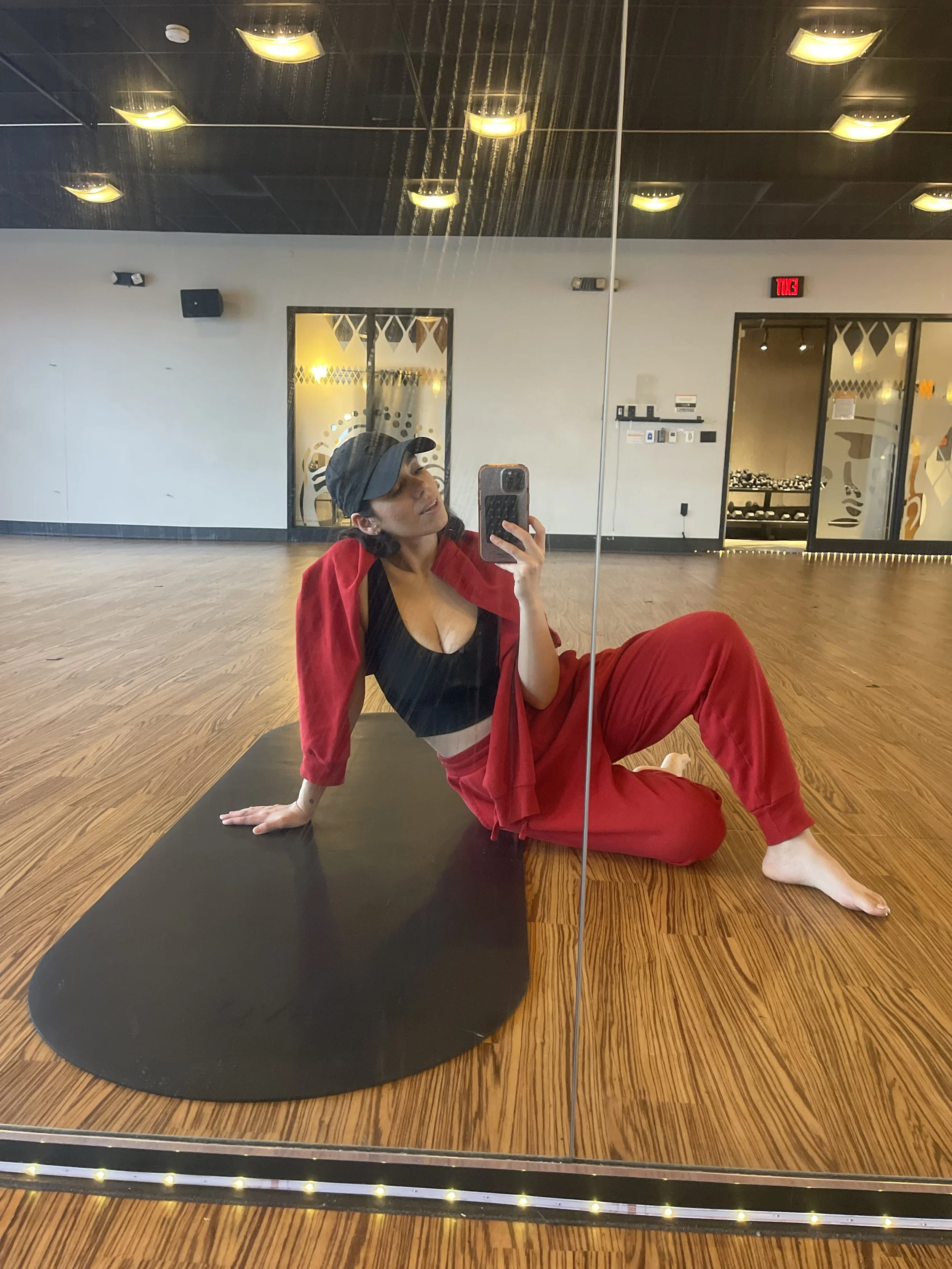 a woman taking a picture of herself in the mirror seated on the floor of a yoga studio with the negro mate capsula mat rolled out next to her.