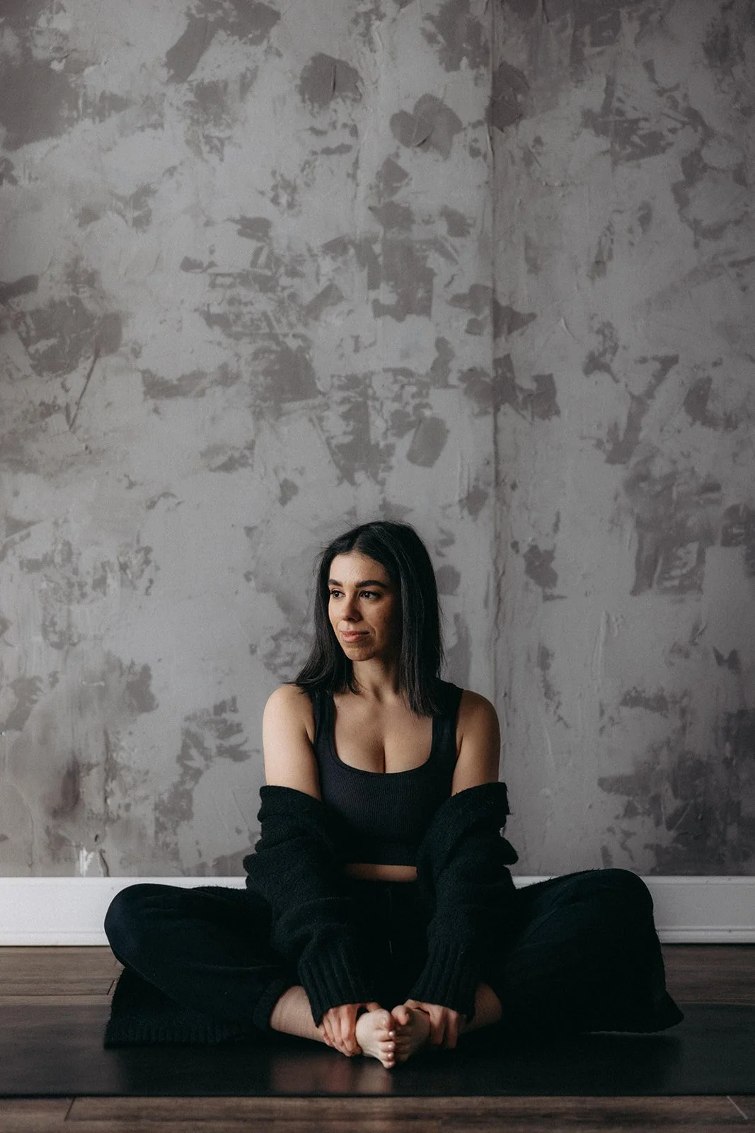 A woman dressed in a black tank top and black jogger pants with a black cardigan off her shoulders seated in a butterfly pose on the Negro Mate Capsula Mat.