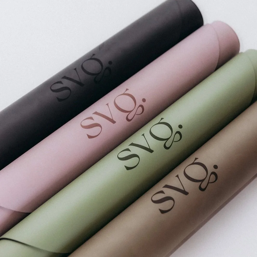 Four rolled-up Capsula Wellness Mats with the brand 'svg.' printed on each in different colors: black, pink, green, and beige.