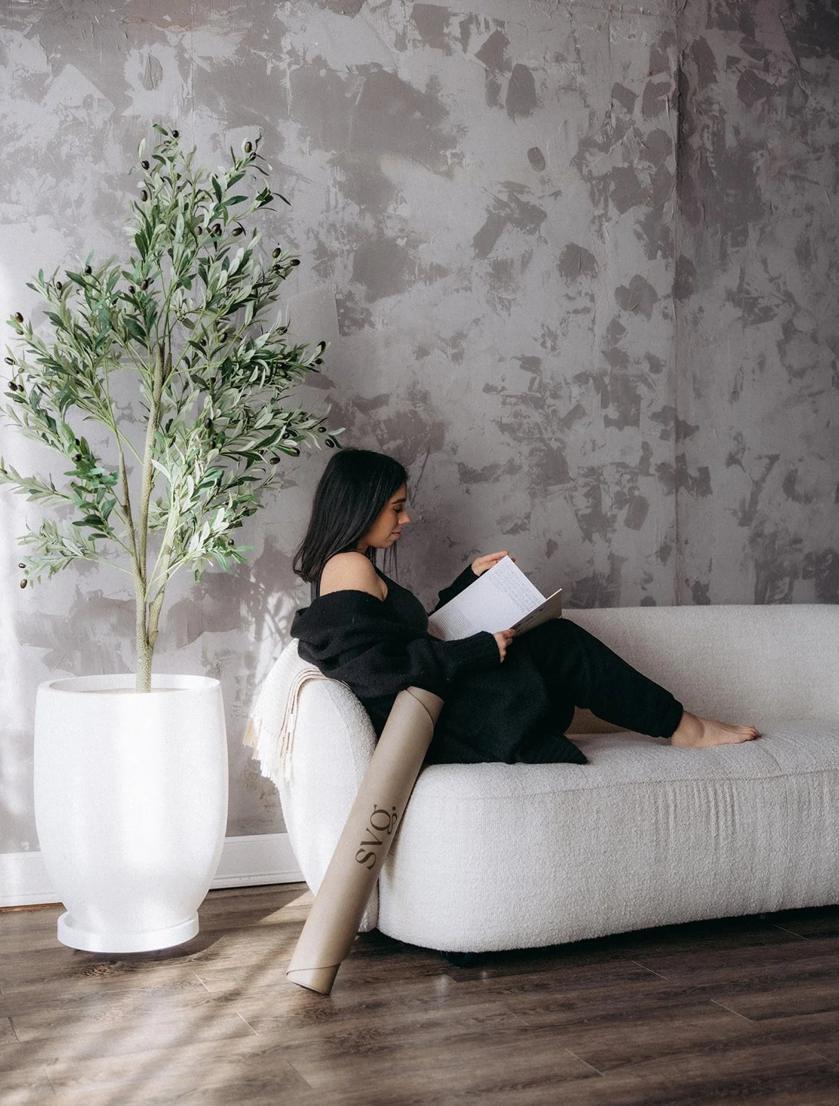 A young woman with dark hair isitting on a light-colored sofa, reading a book. A large potted plant is on her left, and a rolled-up Capsula Wellness Mat leans against the sofa. The background features textured, gray-painted walls and wooden flooring.