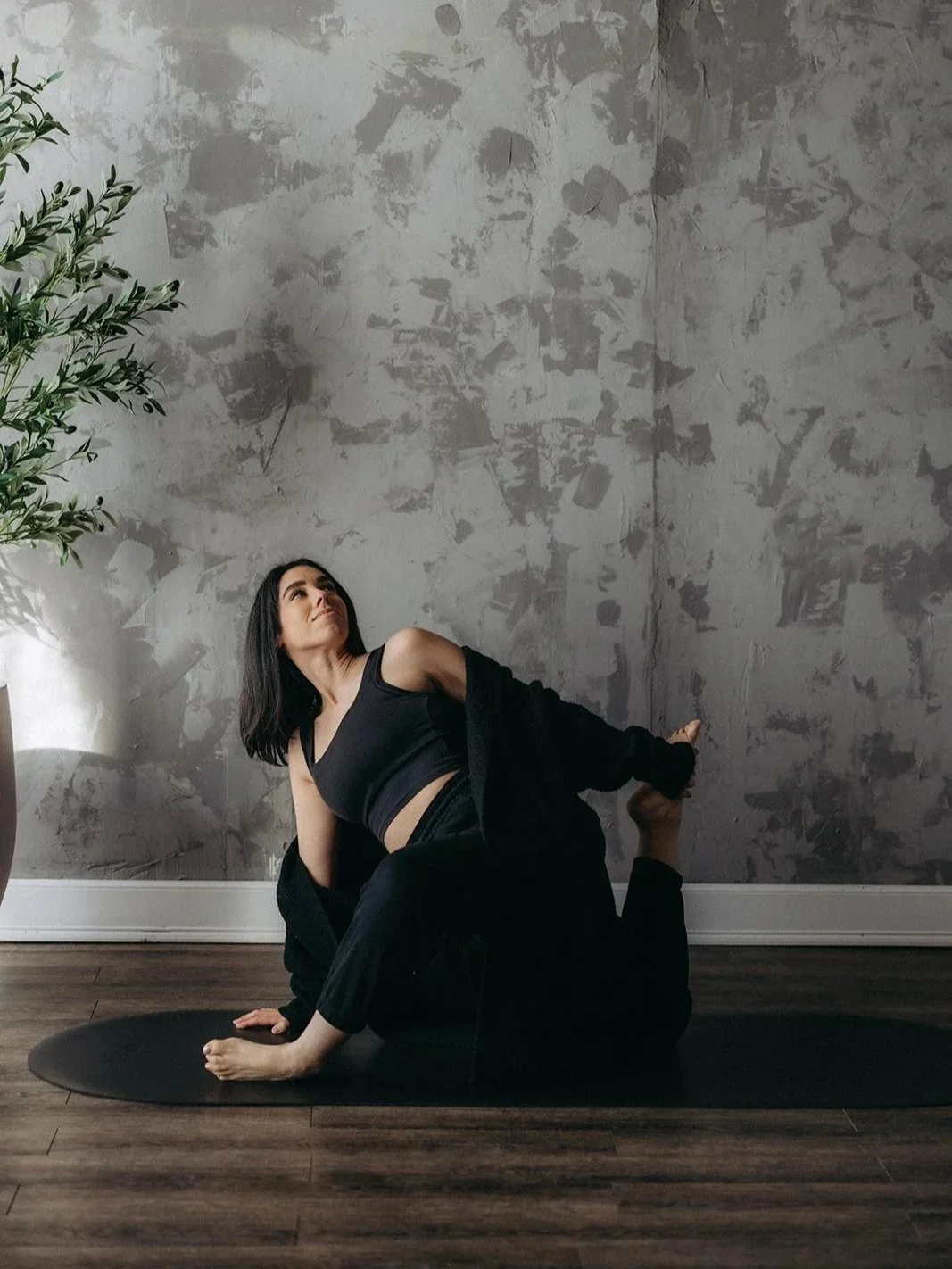 A woman dressed in black joggers, a black tank, and a chunky black knitted cardigan doing a grounded yoga pose on the negro mate capsula mat with a beautiful soft interior design as the backdrop.