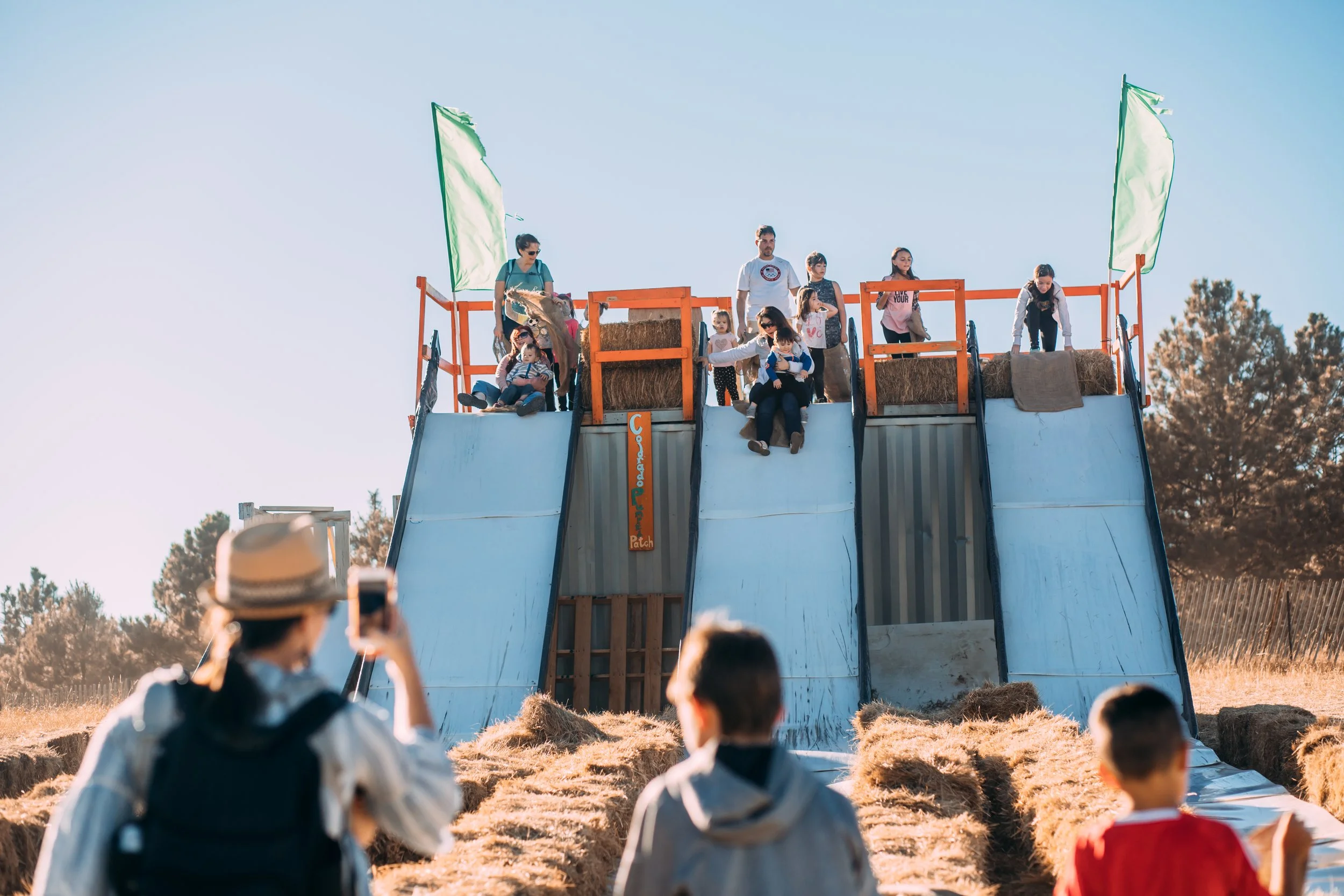 Children and adults descending a large outdoor slide at a fair or festival, with hay bales at the bottom and flags at the top, in a rural setting.