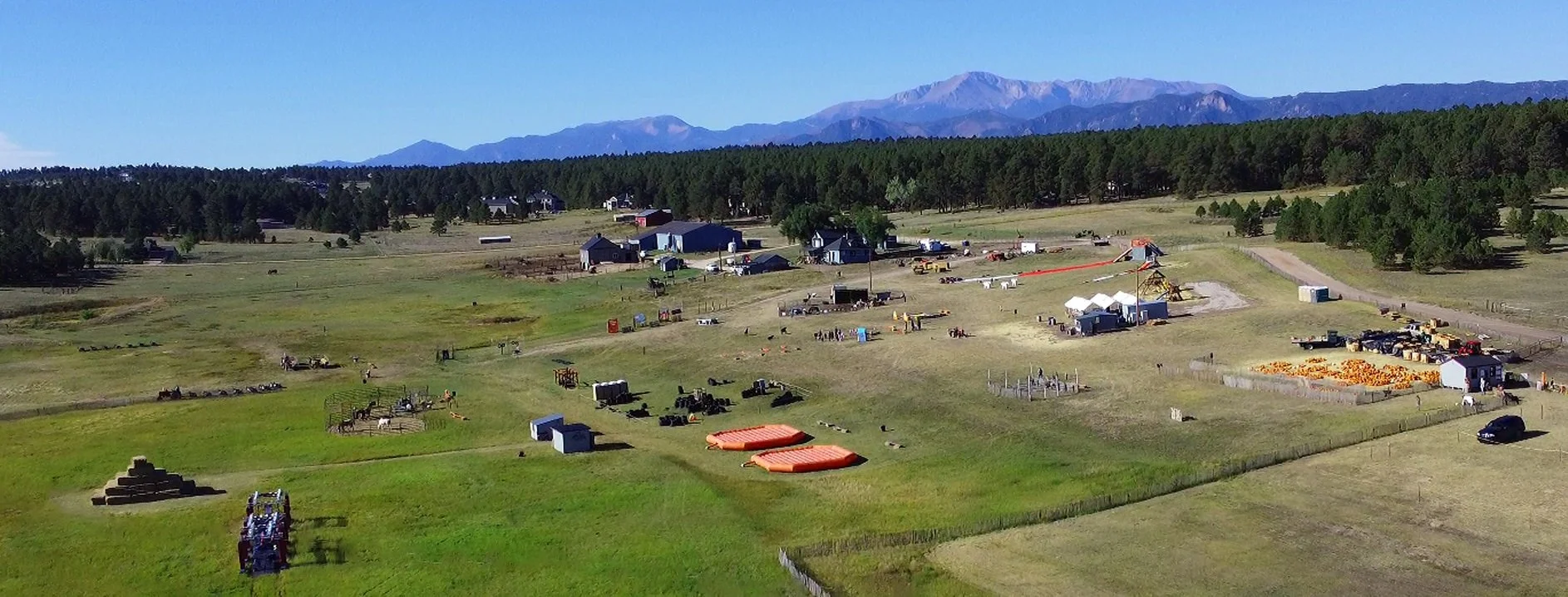 A rural farm with horses, trailers, and pumpkins, surrounded by trees and mountains in the background.
