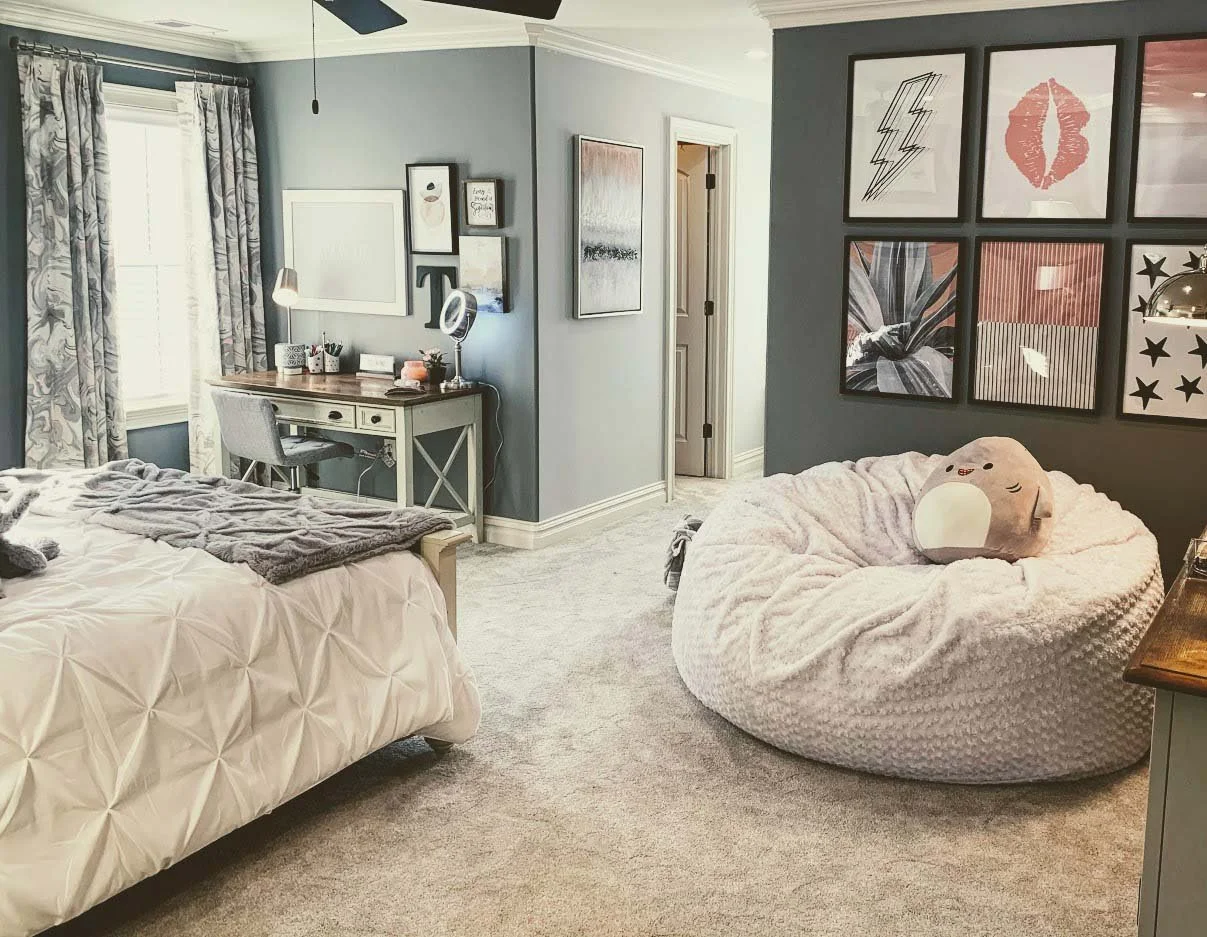 Cozy bedroom with gray walls, a bed with white bedding, a large bean bag chair with a cute plush toy, a wooden desk with a chair and lamp, and framed artwork on the walls.