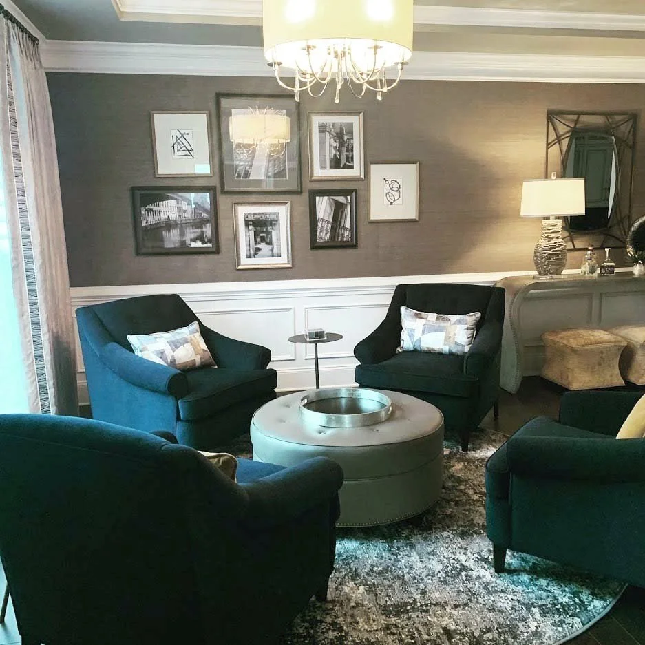 Elegant living room with dark chairs, framed wall art, round ottoman, and chandelier.