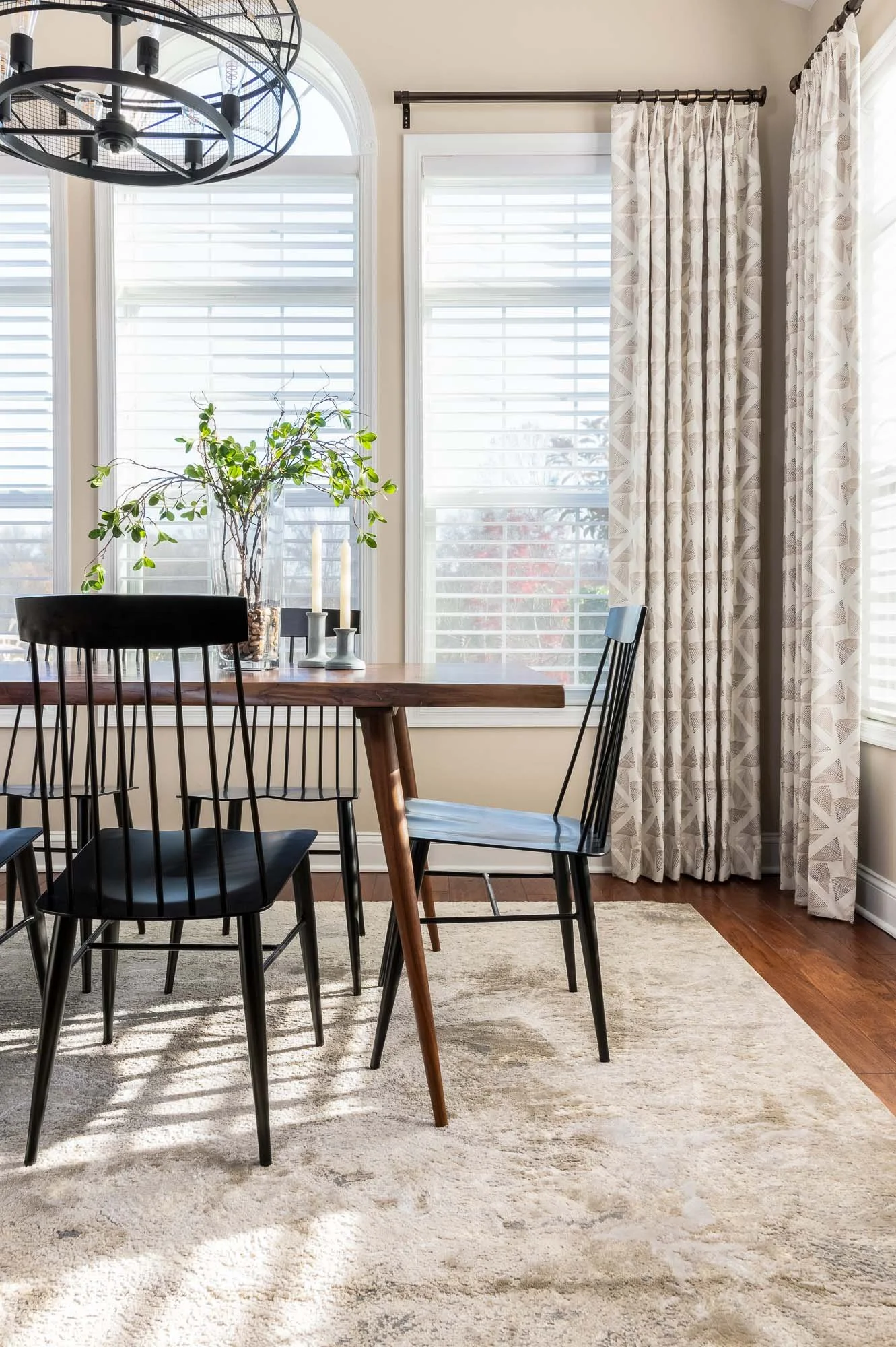 Modern dining room with black chairs, wooden table, houseplant in vase, two white candles, large windows with shutters, patterned curtains, and chandelier.
