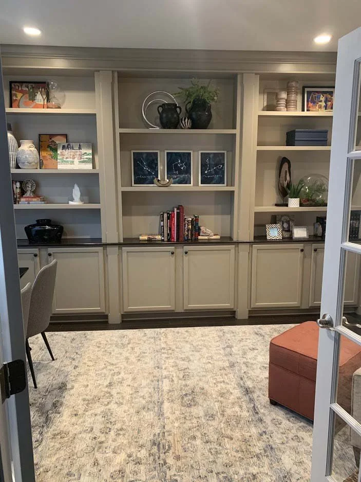 Interior of a home office with built-in shelves displaying books, vases, and decorative items. There is a cushioned ottoman, a patterned rug, and a partial view of a chair. The room is well-lit and neatly organized.