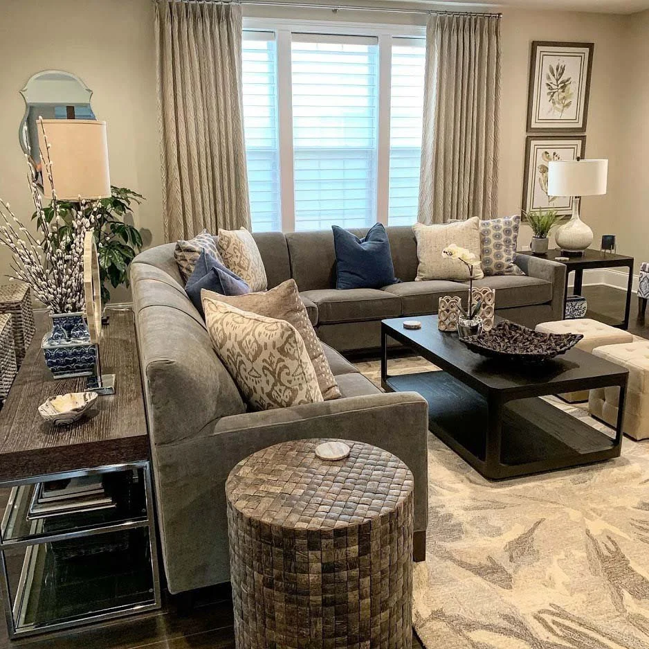 Modern living room with a gray sectional sofa, decorative pillows, wooden coffee table, lamps, and wall art. Large windows with curtains.