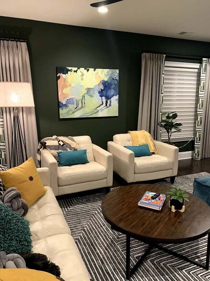 Cozy living room with white leather chairs, teal and yellow pillows, abstract art on a green wall, wooden coffee table with plant and magazines, patterned rug, and large windows with drapes.