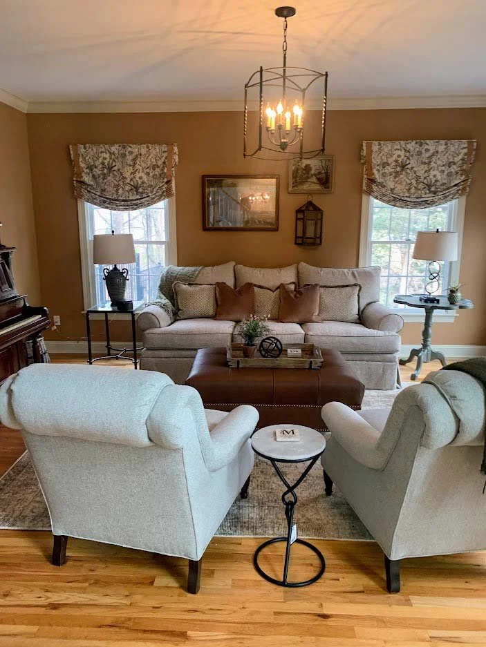 Elegant living room with beige walls, a light gray sofa, two armchairs, a leather coffee table, decorative pillows, a chandelier, and framed wall art.