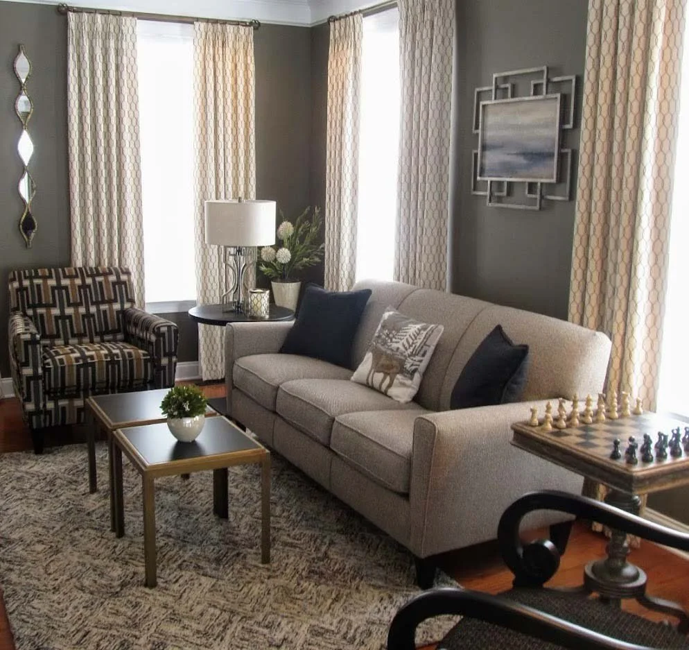 Modern living room with beige sofa, patterned armchair, and chess table on a rug. Curtains frame the windows, and a painting hangs on a gray wall.