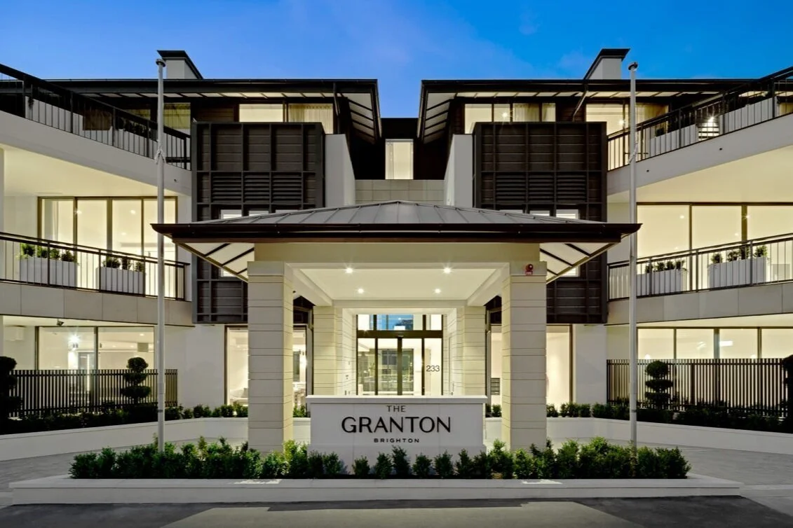 Exterior of The Granton building at dusk with modern architecture and large windows, entrance lit up, surrounded by lush landscaping.