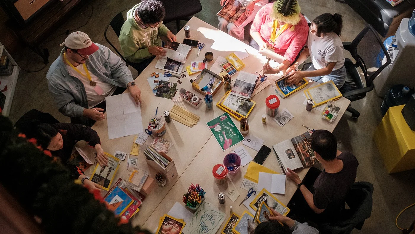 bird's eye view of a wooden table with people sitting around it flipping through magazines, finding images to collage with. the table also has pencils, markers, tapes and zine templates to create with.