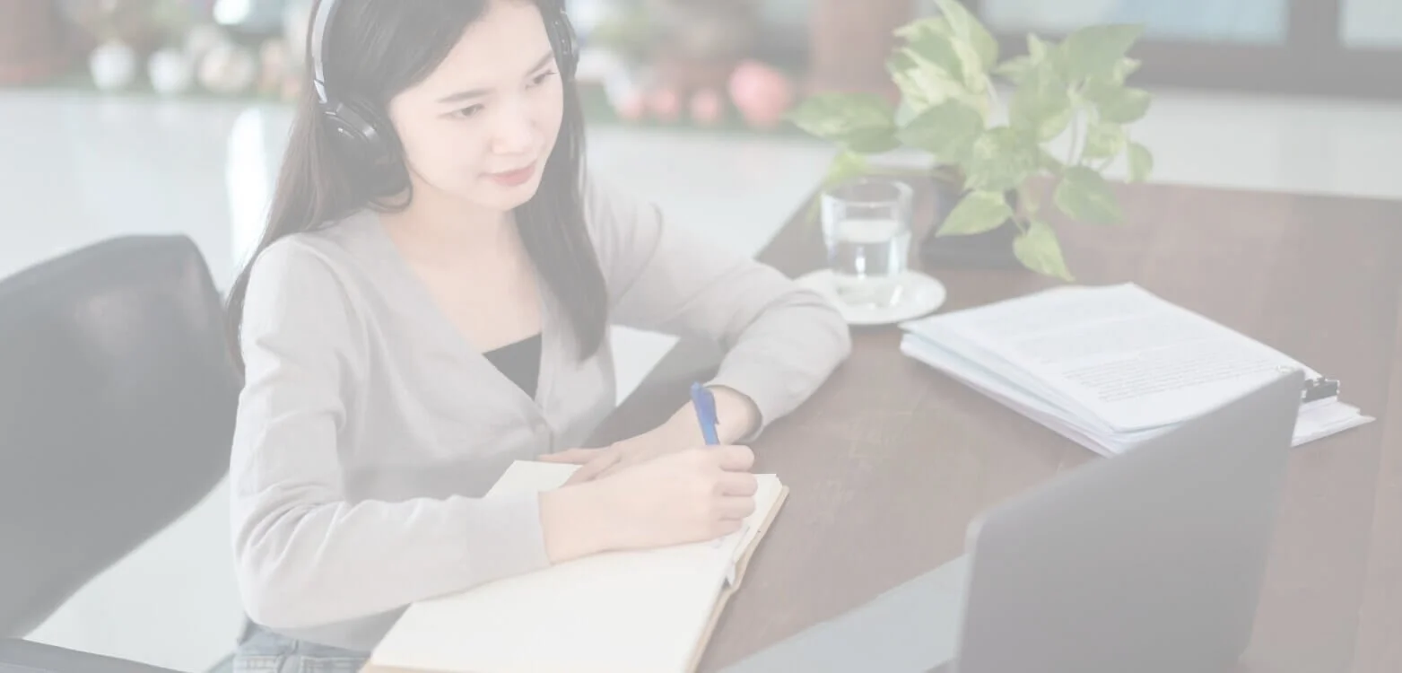 A woman wearing headphones, taking notes with a pen, sitting at a desk with a laptop, and a glass of water, with a plant and documents in the background.