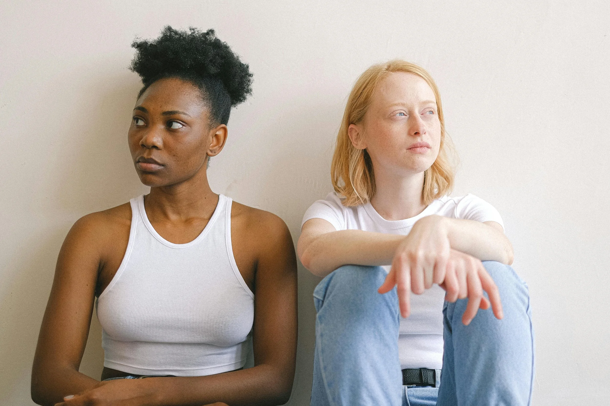 Two women sitting on the floor against a plain wall, looking away from each other in a quiet indoor setting.