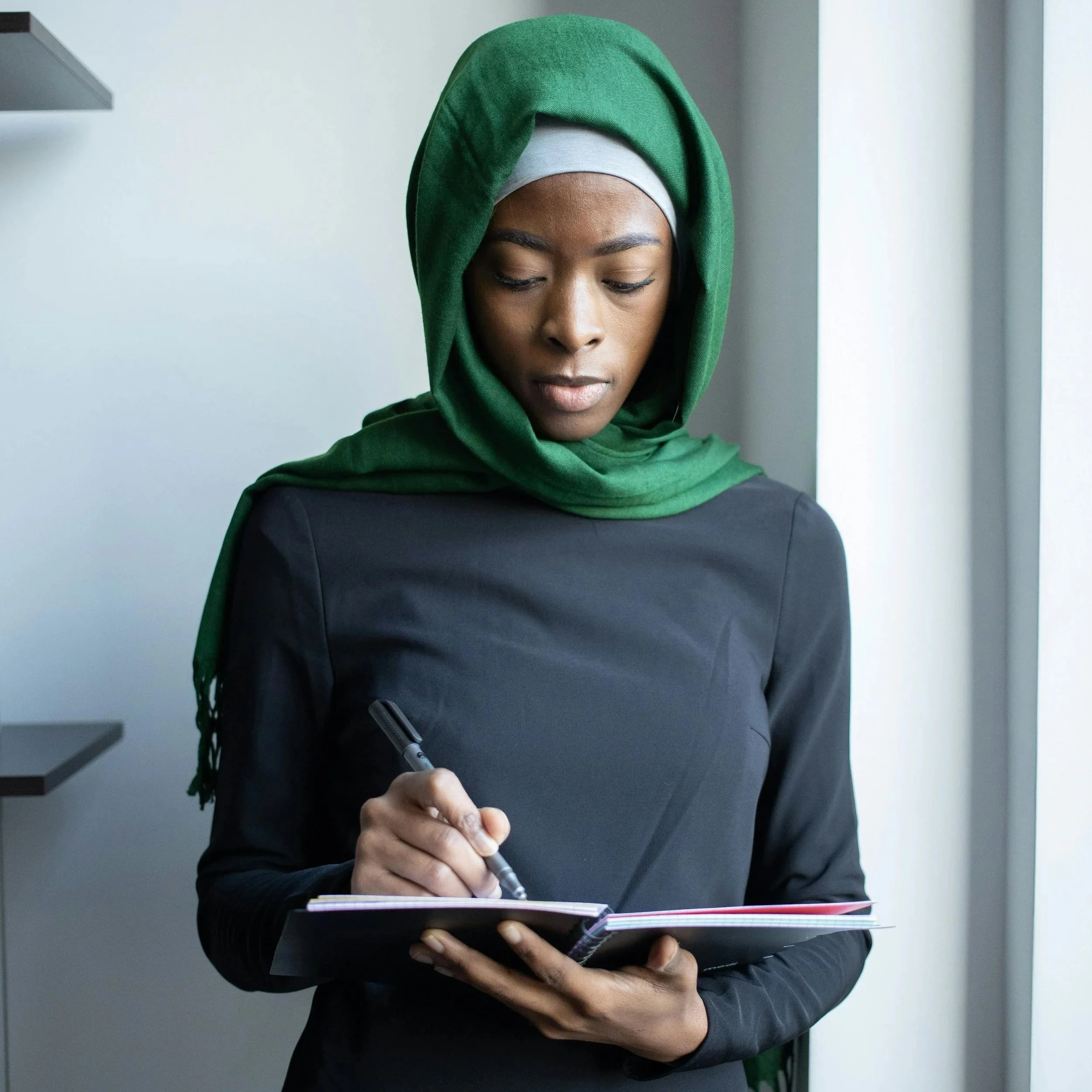 Woman standing near a window writing goals and ideas in a notebook while planning her next steps.