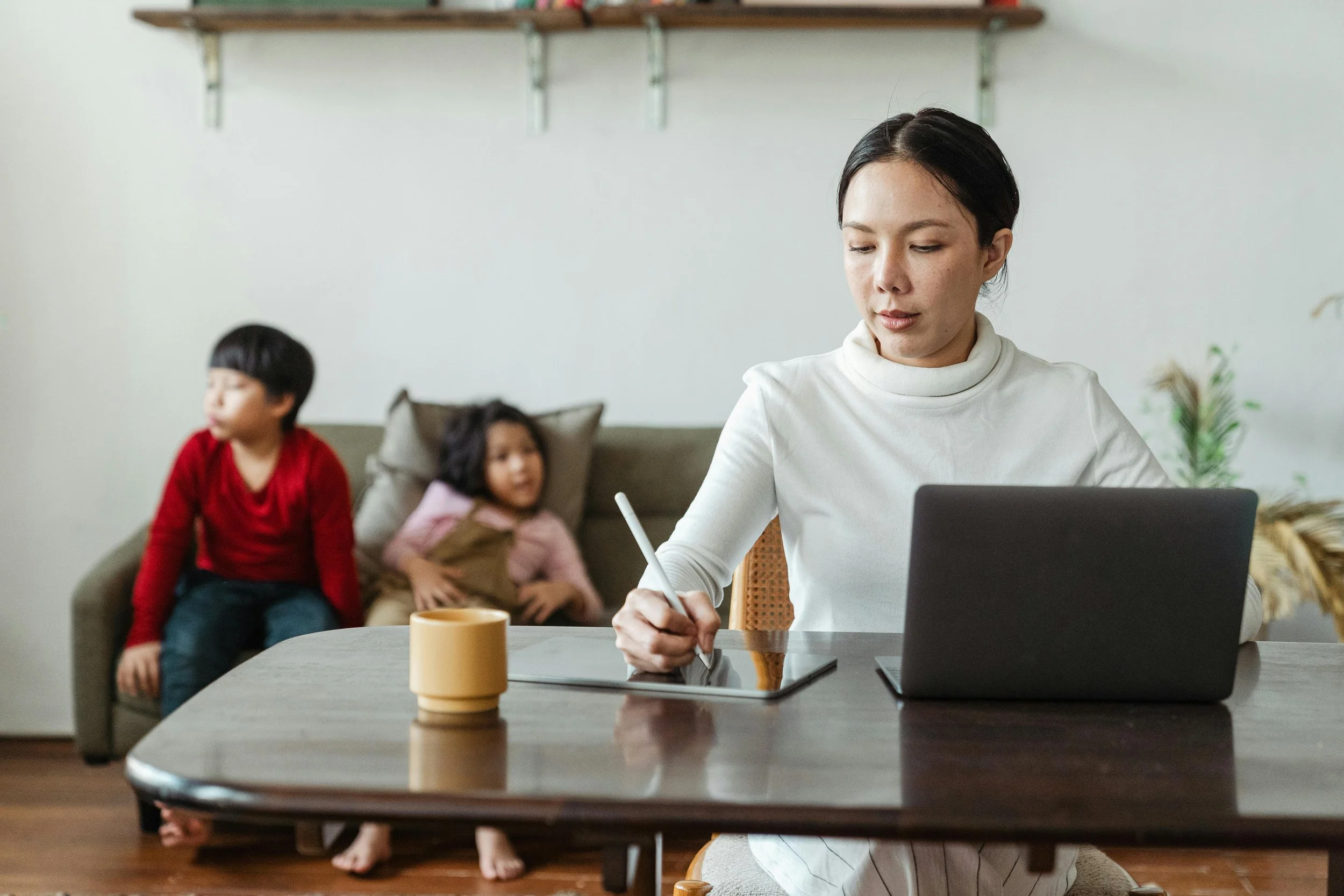 Woman working on a laptop at a dining table while two children sit behind her on a couch, representing the balance of responsibility, distraction, and the quiet pressures that can delay personal change.