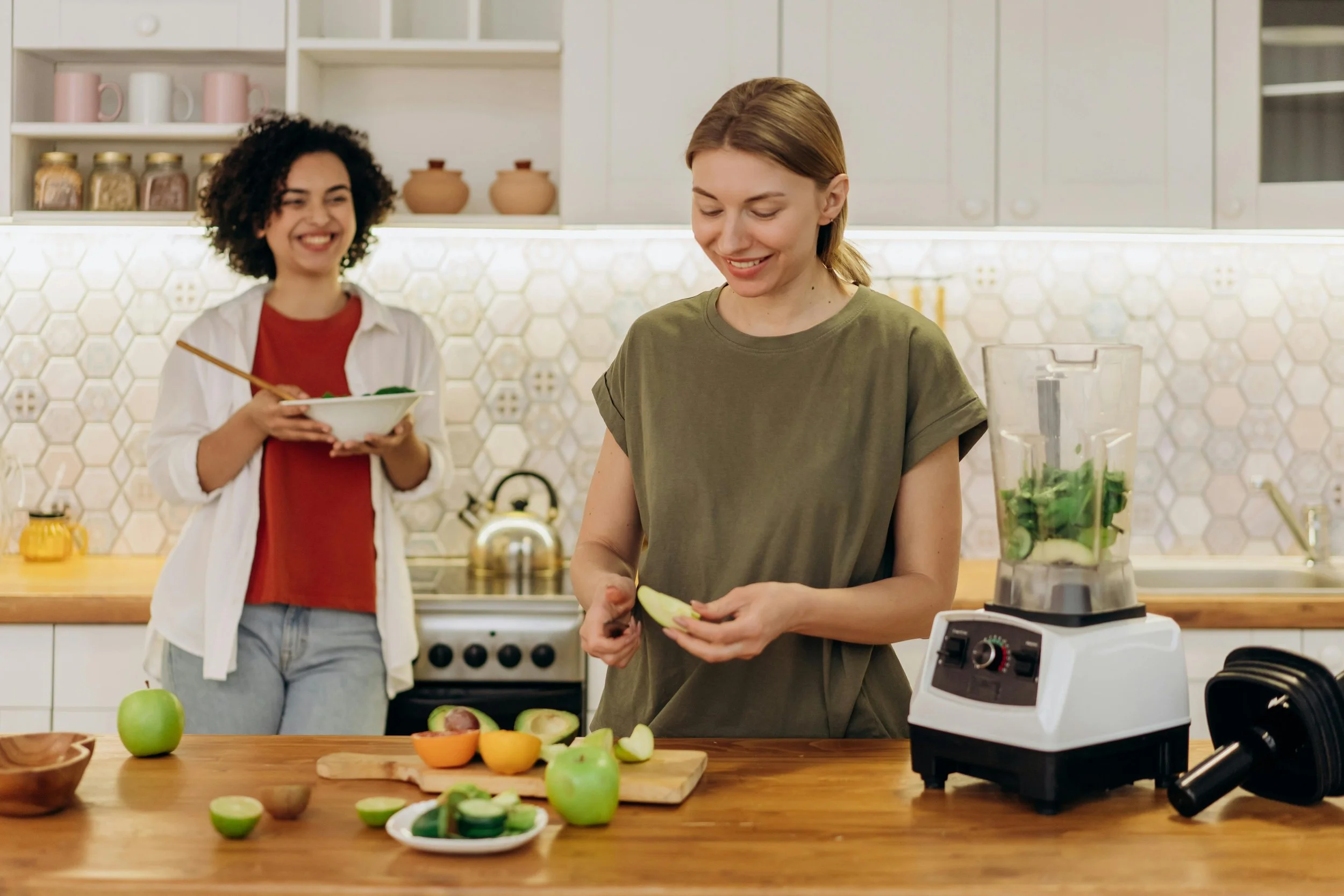 Two people preparing food in a kitchen, with one chopping fruit at a counter and another standing nearby holding a bowl.