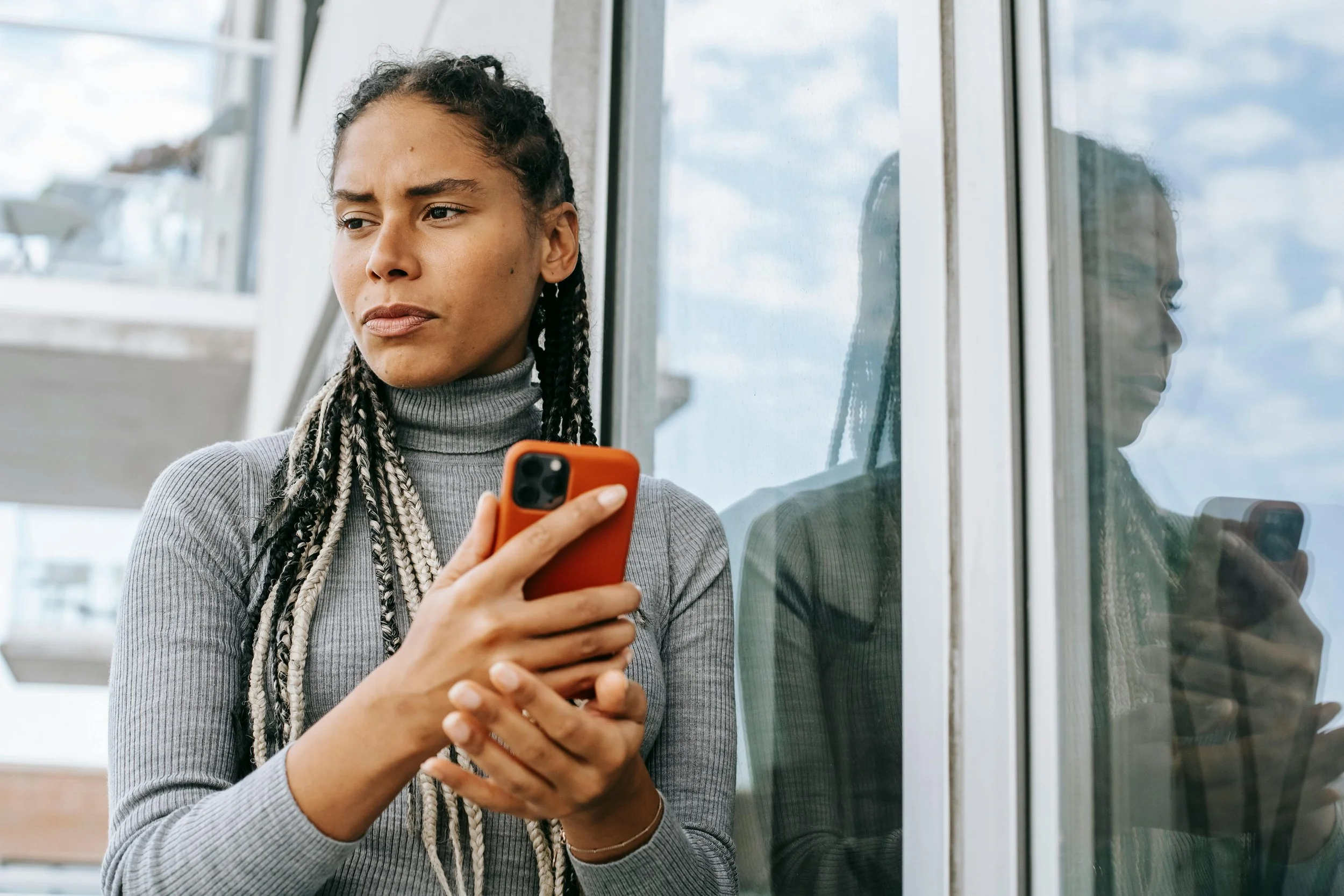 Woman standing by a window holding her phone and looking outside with a serious expression, symbolizing contemplation, decision making, and the quiet tension before making a life change.