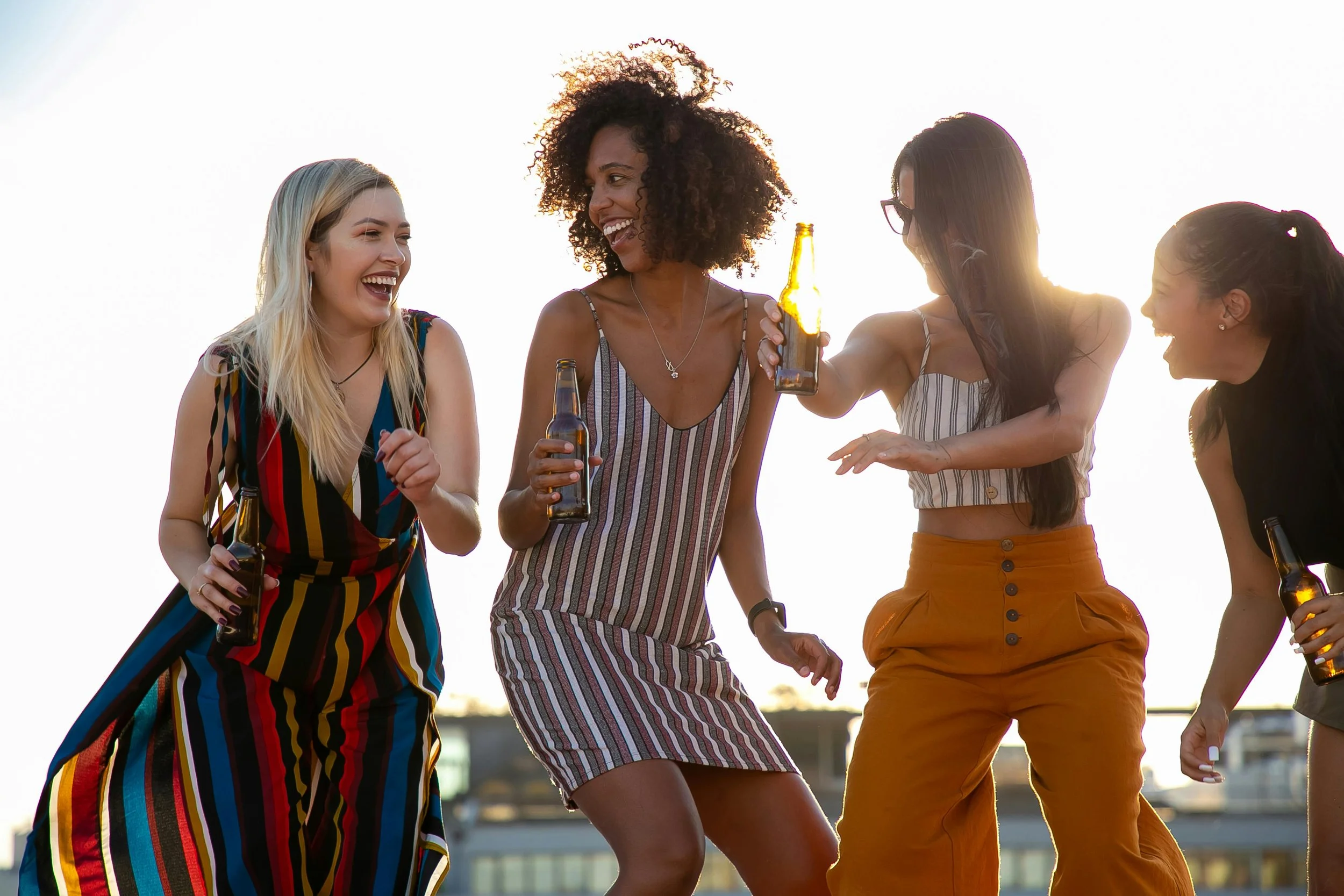 Group of women standing outdoors at sunset, laughing and holding drinks together in a relaxed social setting.