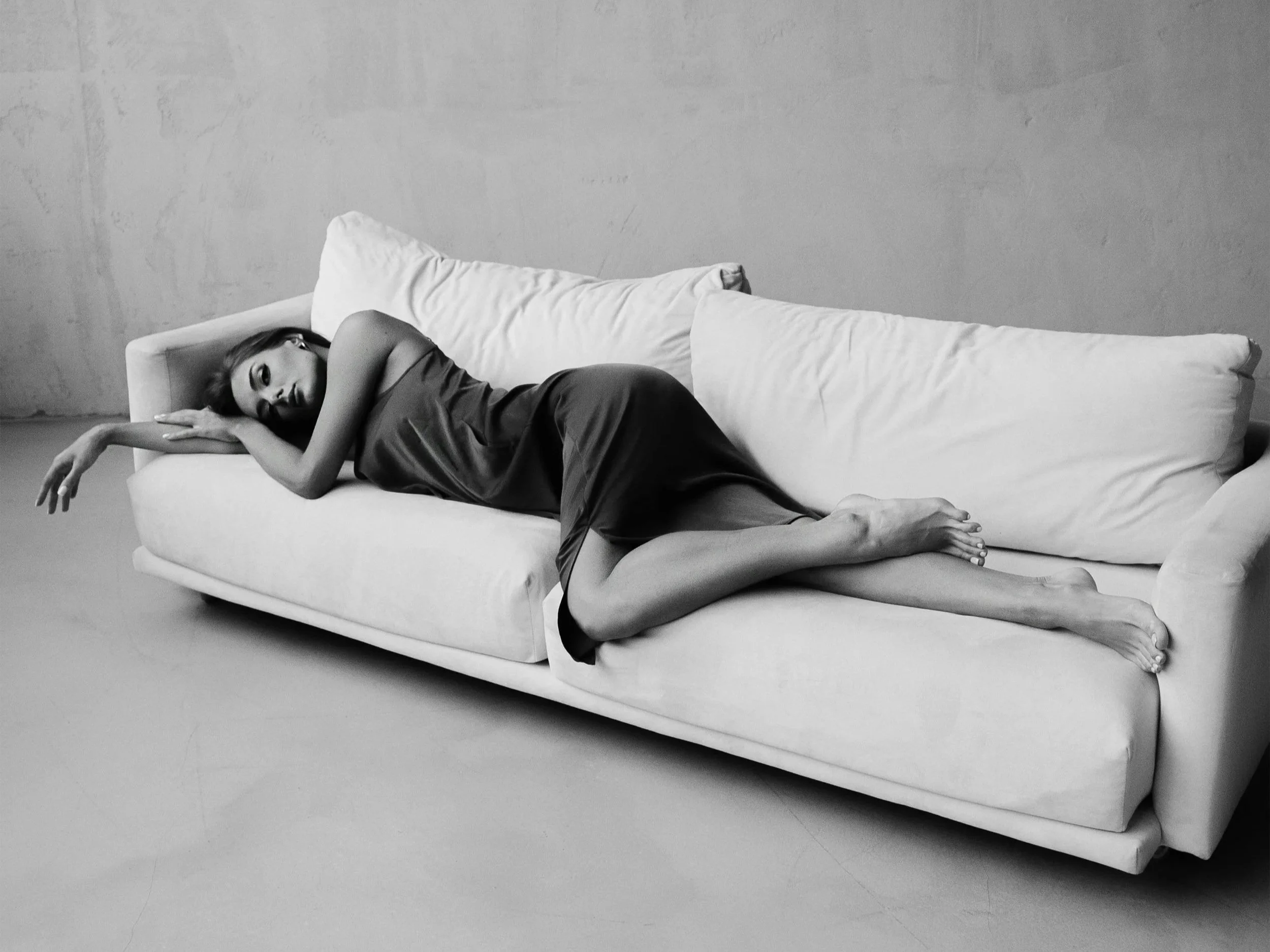 Woman lying alone on a sofa in a minimalist room, appearing distant and contemplative, symbolizing emotional exhaustion, stillness, and the quiet weight of staying in a life that no longer feels aligned.