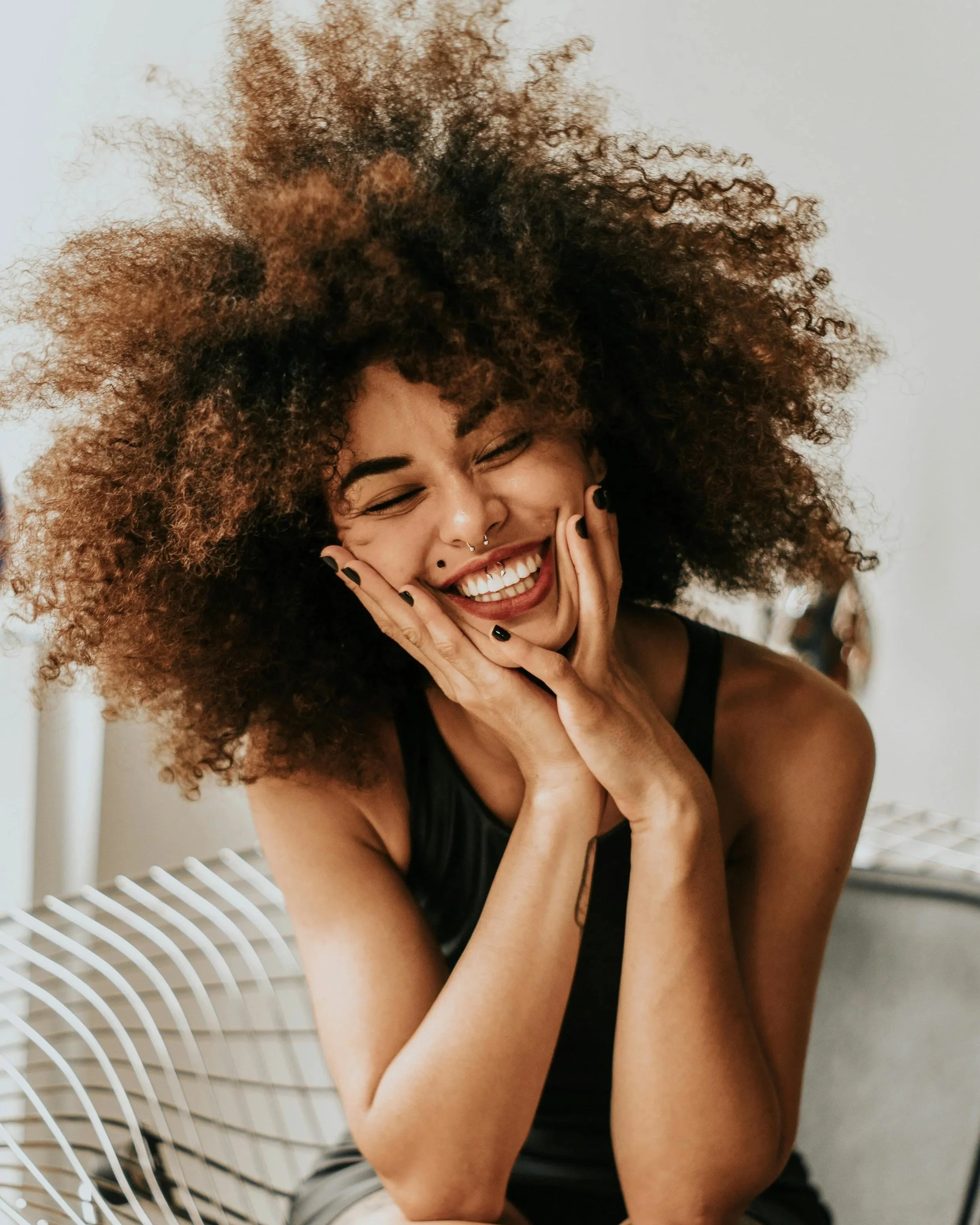 Smiling woman with natural curly hair sitting in a modern chair, representing confidence, balance, and joyful productivity.