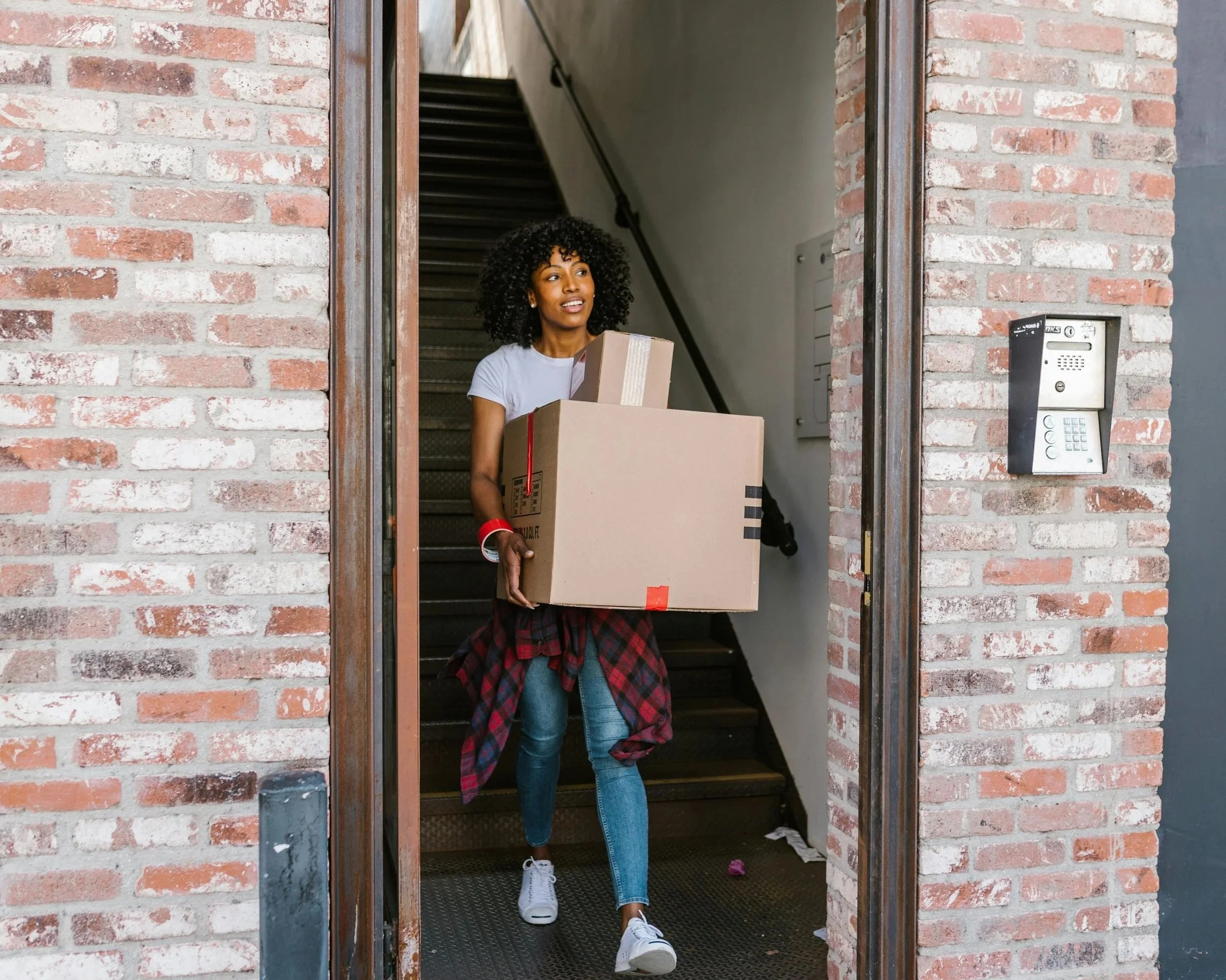 Woman carrying cardboard moving boxes out of a brick apartment building, symbolizing transition, independence, and stepping into a new chapter of life.