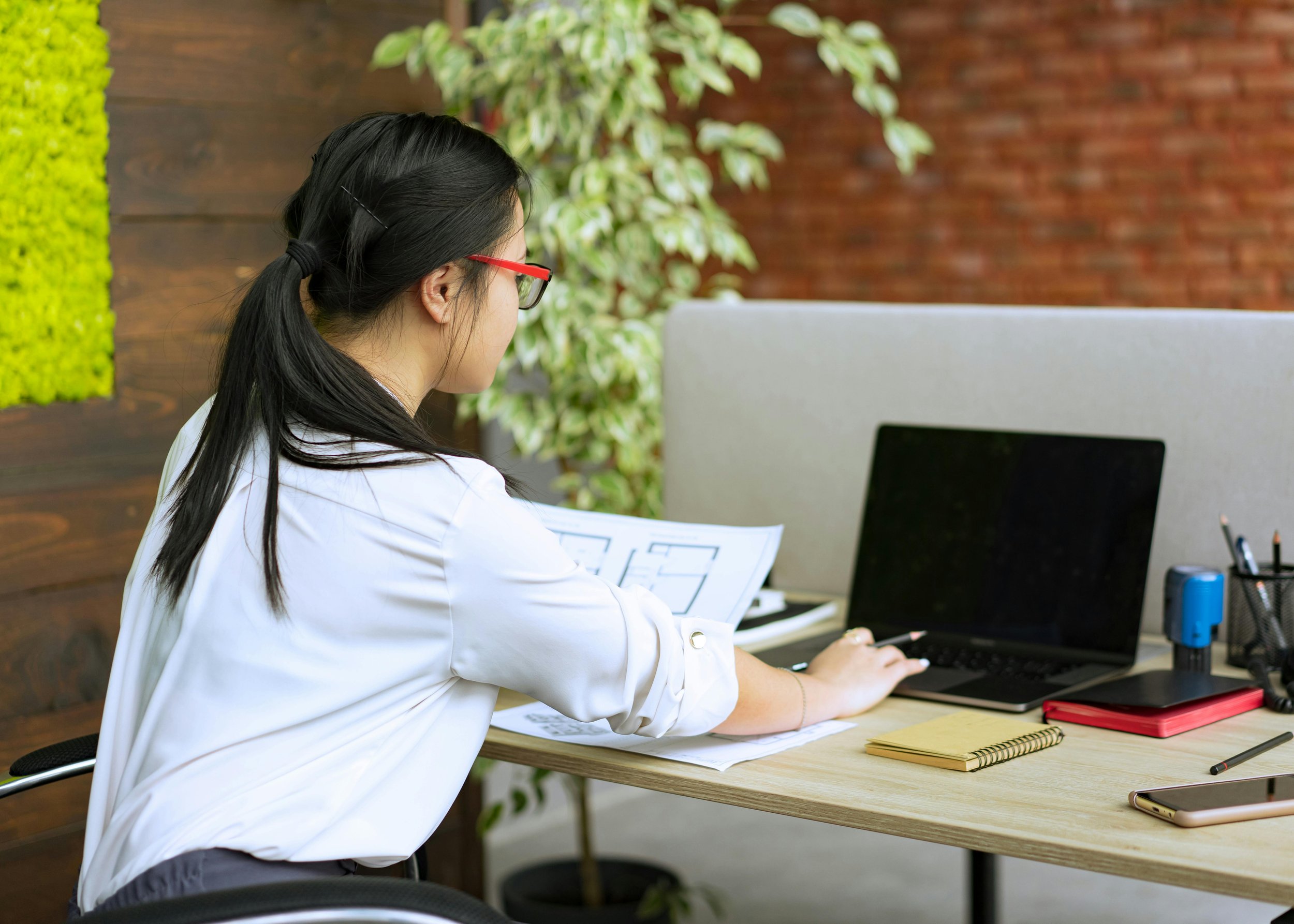 A woman sits at a desk working on her laptop, surrounded by work materials, appearing focused and productive while managing multiple responsibilities.