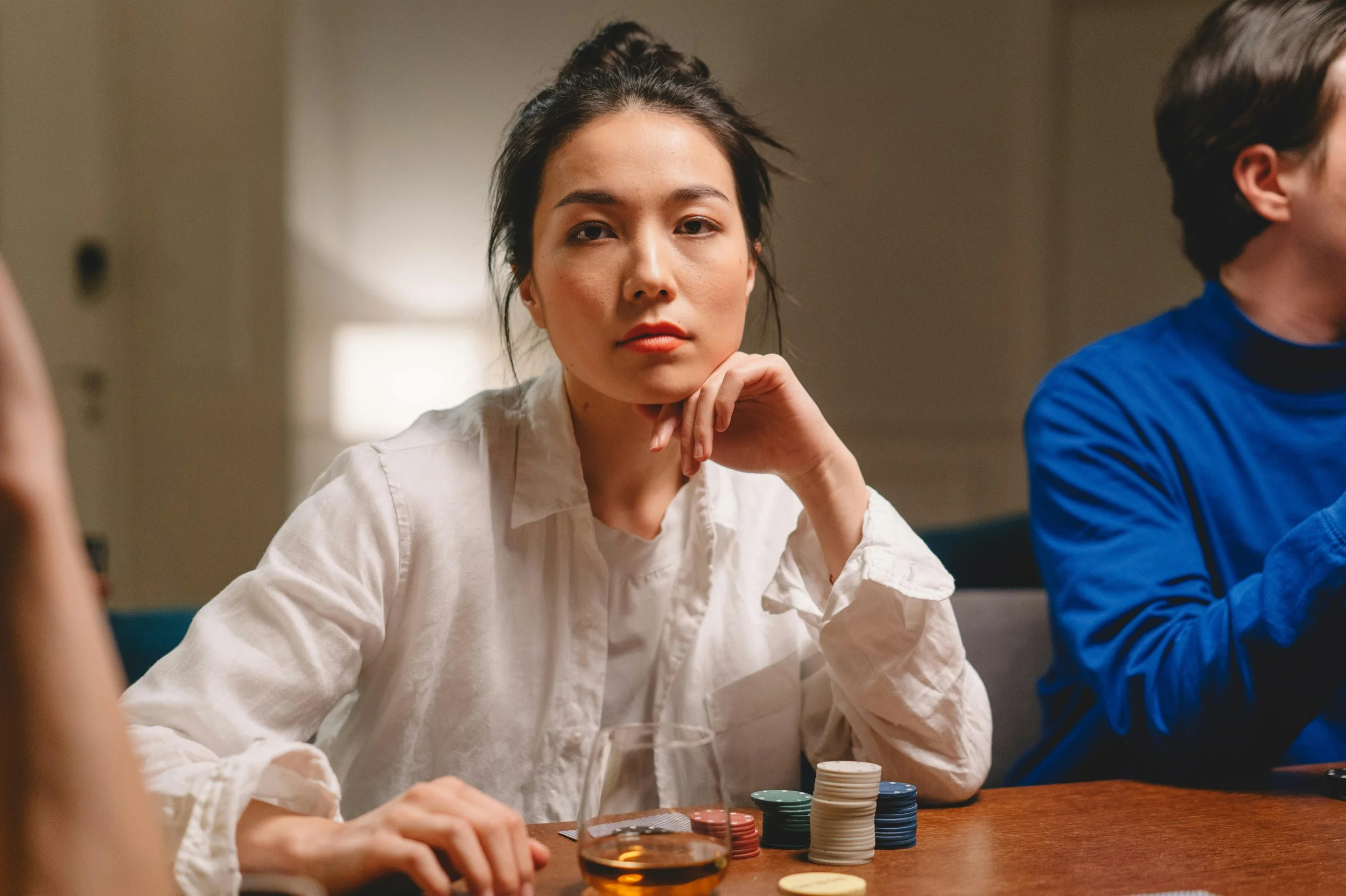 Woman sitting at a table with poker chips and a glass, resting her chin on her hand, appearing calm and reflective in a quiet indoor setting.