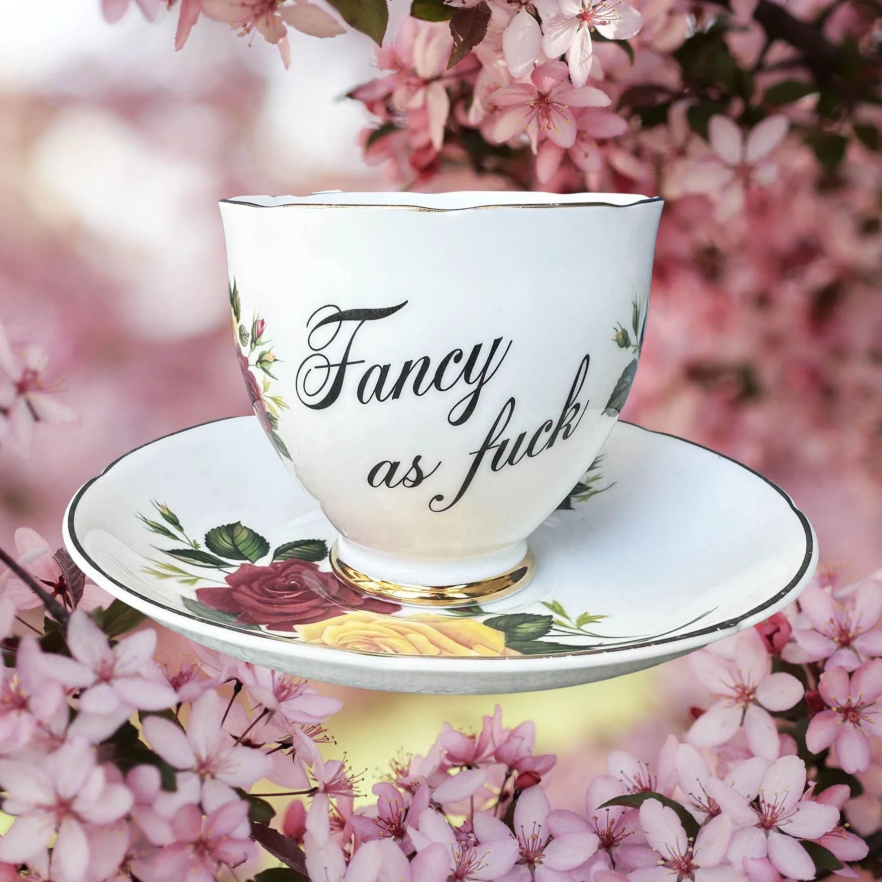 A vintage tea cup and saucer with a red and yellowfloral design, with the words "fancy as fuck" in black cursive writing