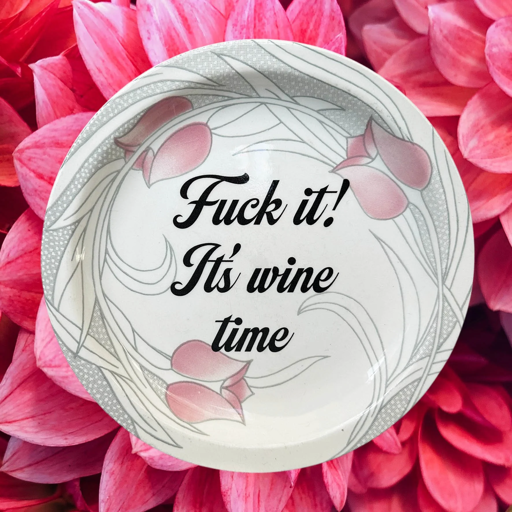 A vintage decorative wall plate with a pink floral design and the words “fuck it! It’s wine time” in black writing
